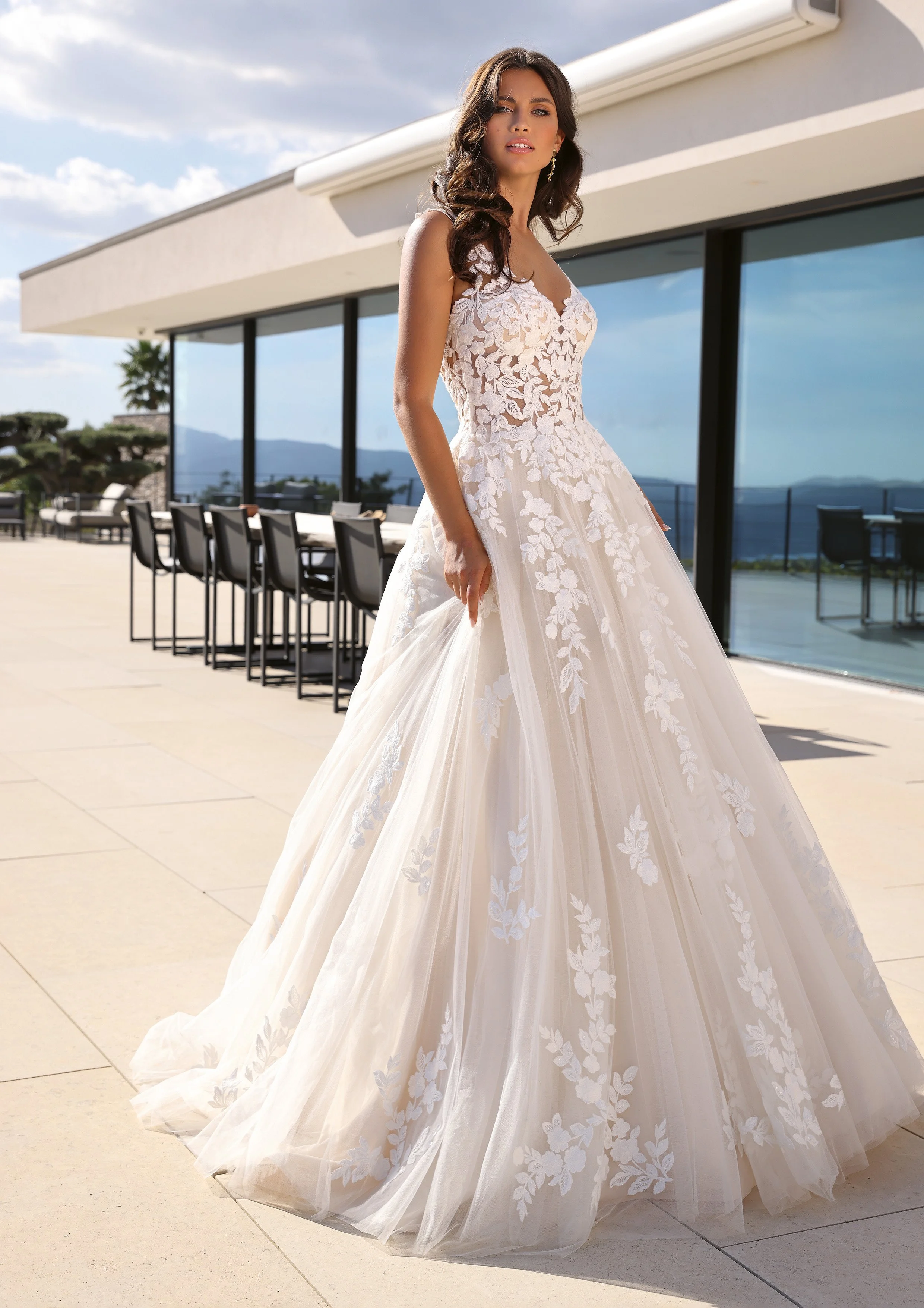 Mulher com vestido de casamento com detalhes de floral e uma saia longa, em um local ao ar livre com varanda e vista para montanhas ao fundo.