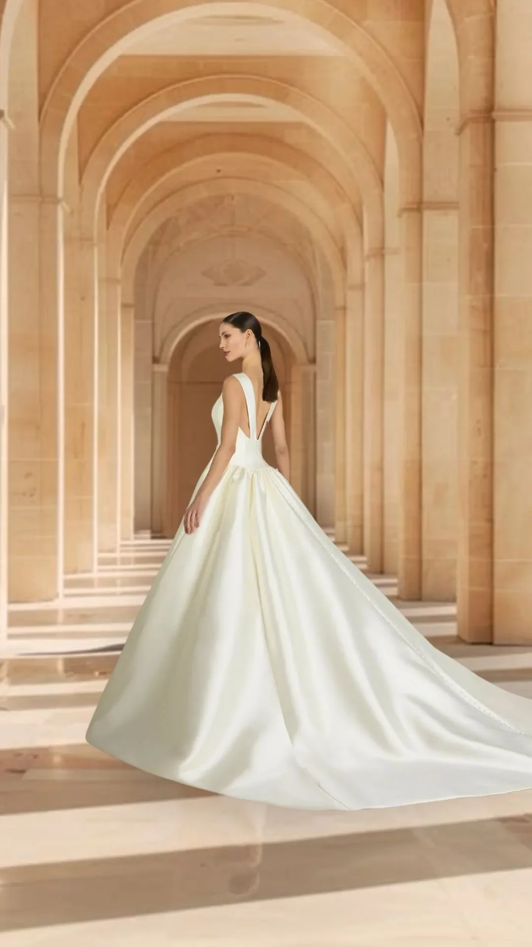 Mulher em vestido de noiva branco de cauda longa, de cabelo escuro e lisasso, de perfil, dentro de um corredor com arcos altos de pedra