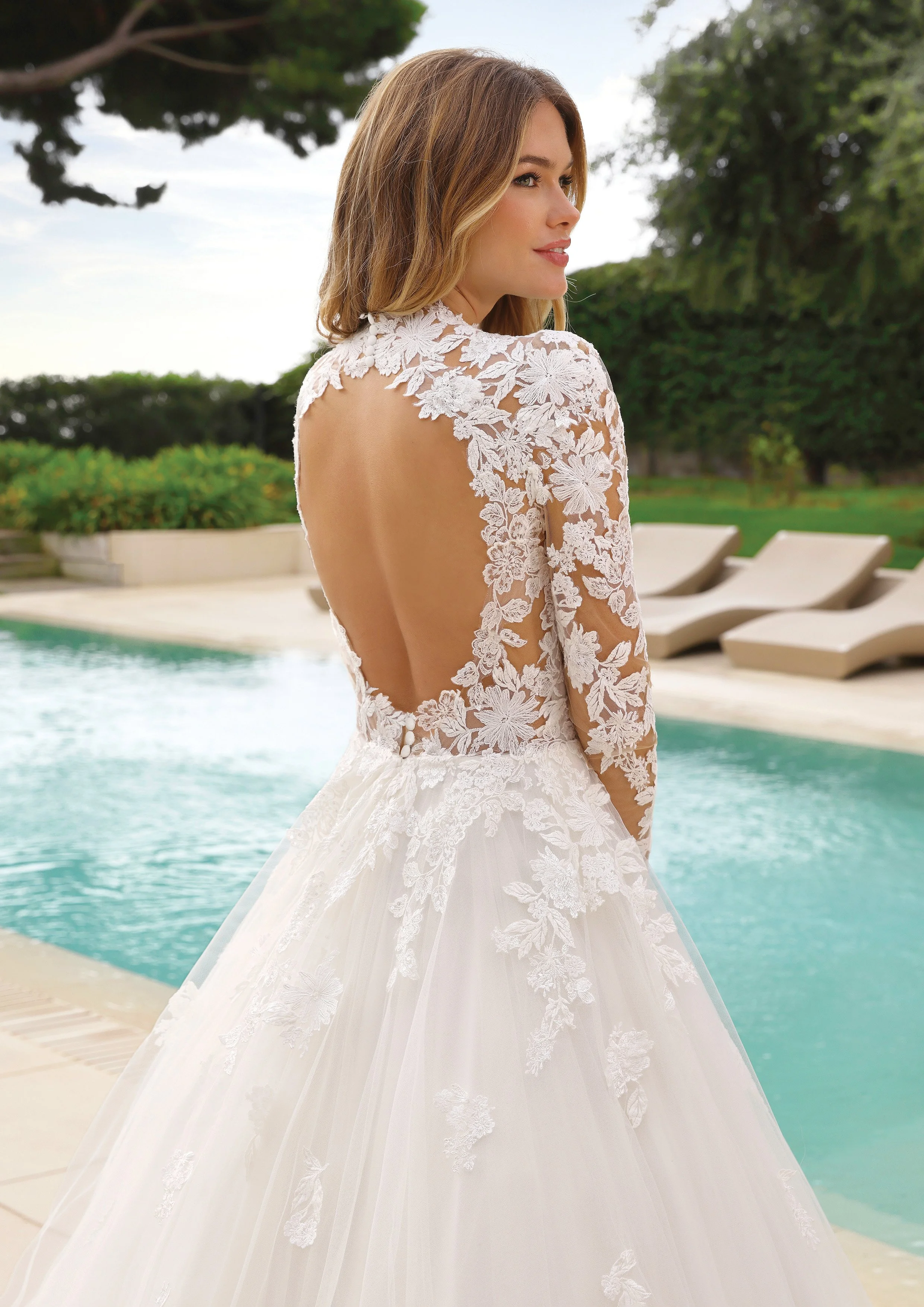 Mulher vestida com vestido de casamento com detalhes de renda, de cabelo castanho, com fundo de piscina e área verde ao ar livre.