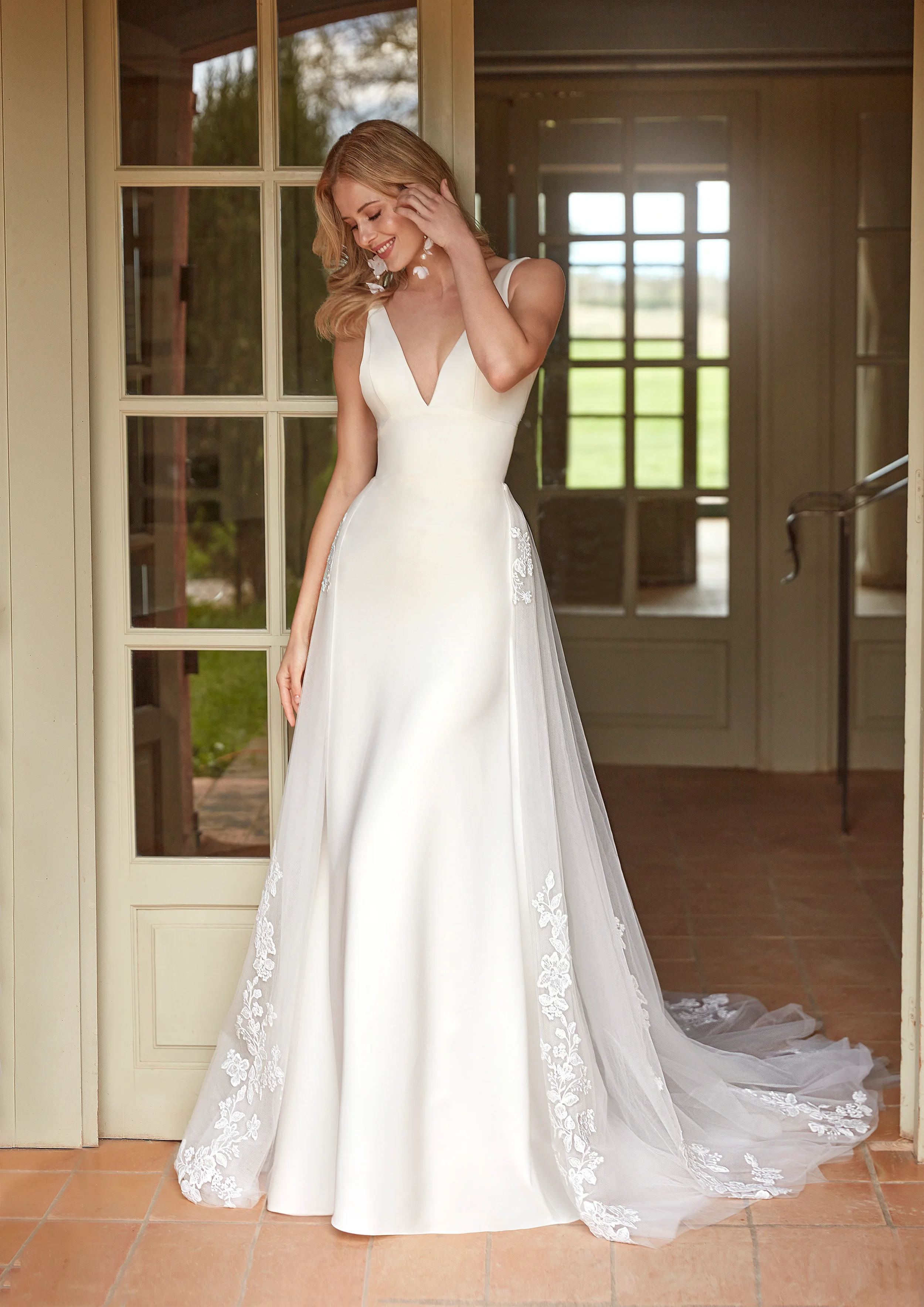 Mulher em vestido de casamento branco com detalhes em renda, sorrindo e ajustando o cabelo, dentro de uma casa com portas de vidro e janelas ao fundo.