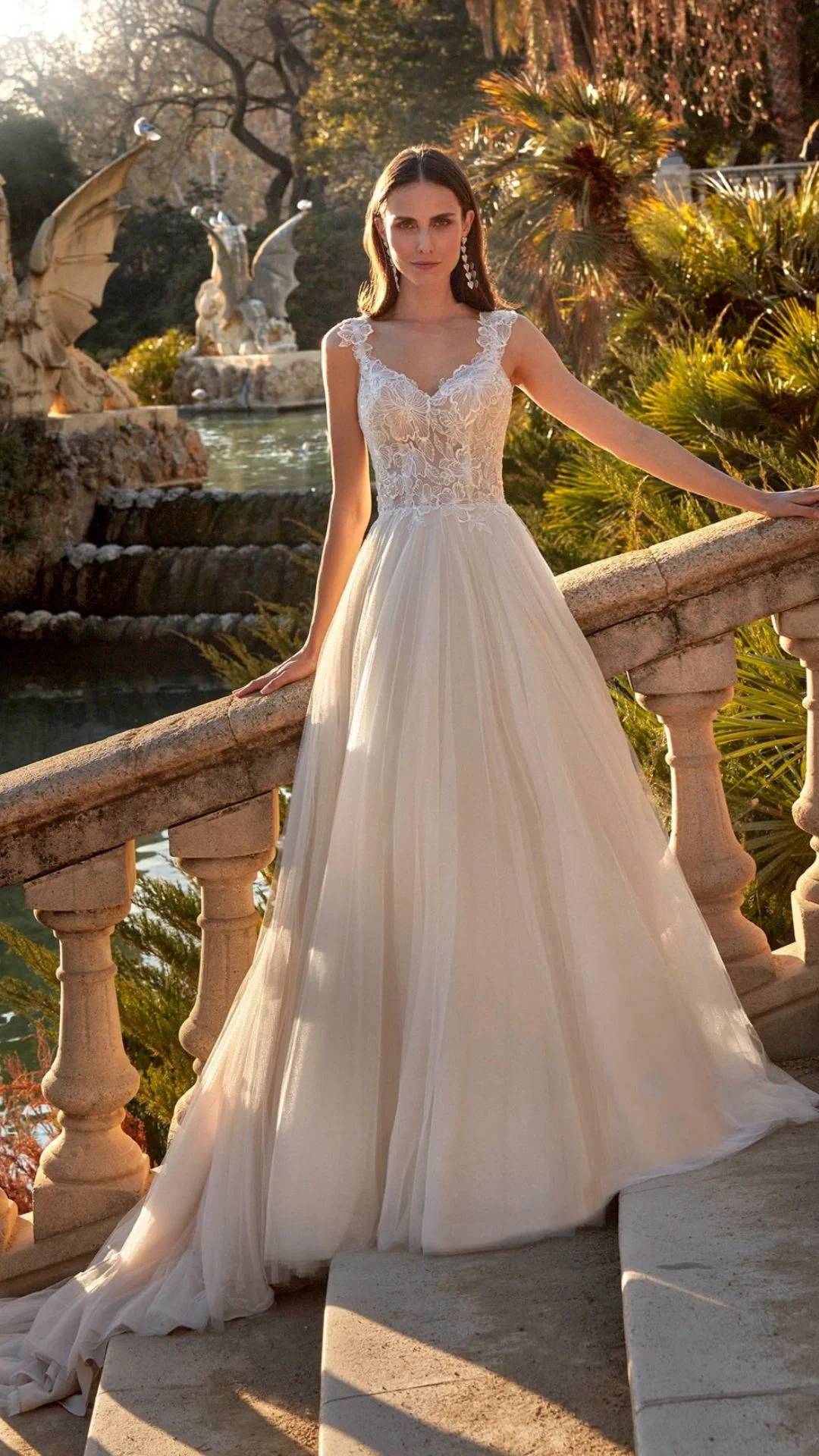 Mestre com vestido de noiva branco e renda, posando ao lado de um jardim com fonte e esculturas de dragões, durante o pôr do sol.