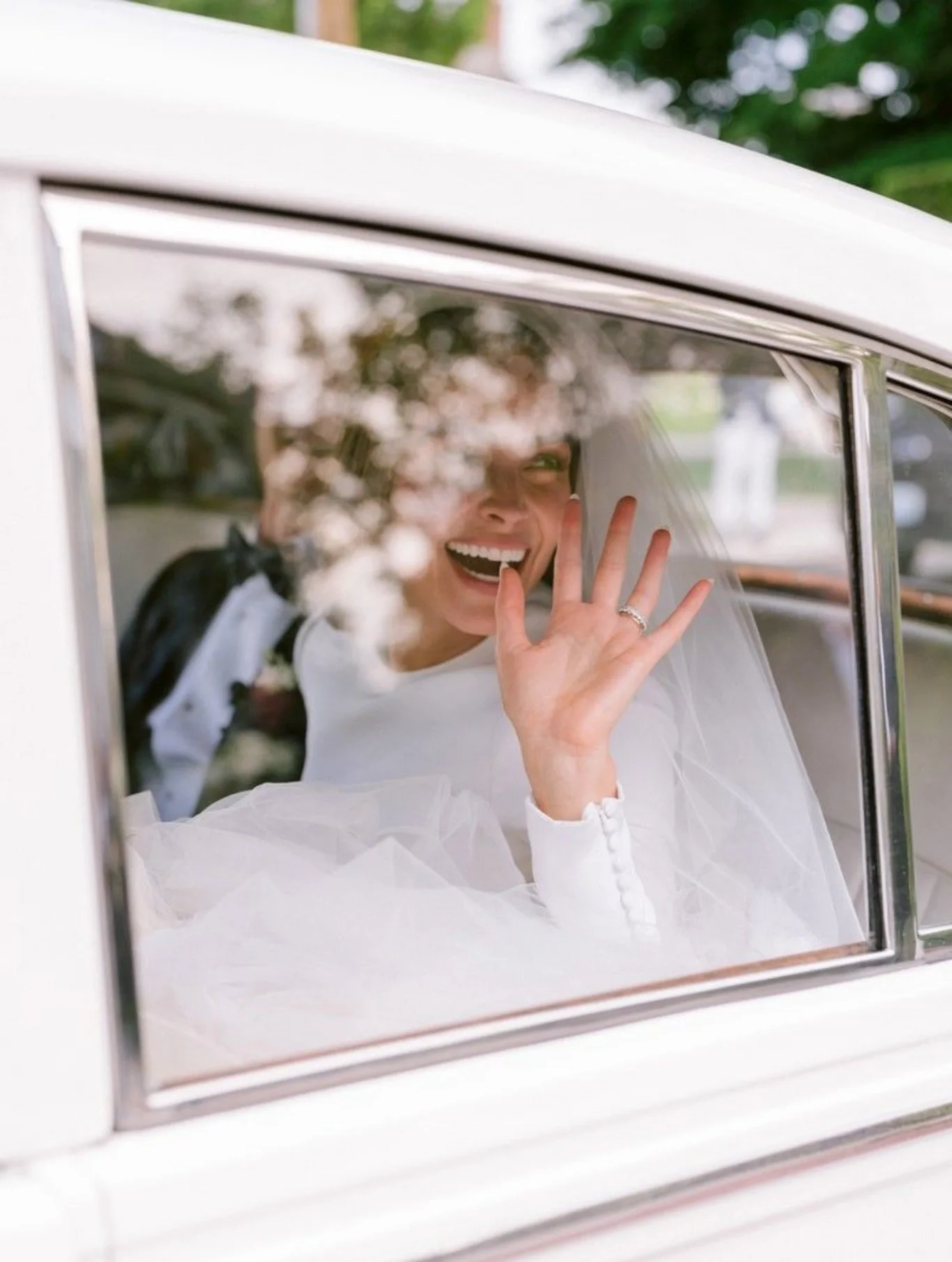 No carro, uma noiva sorridente e acenando pela janela com véu branco, vestido branco e fundo de árvores.