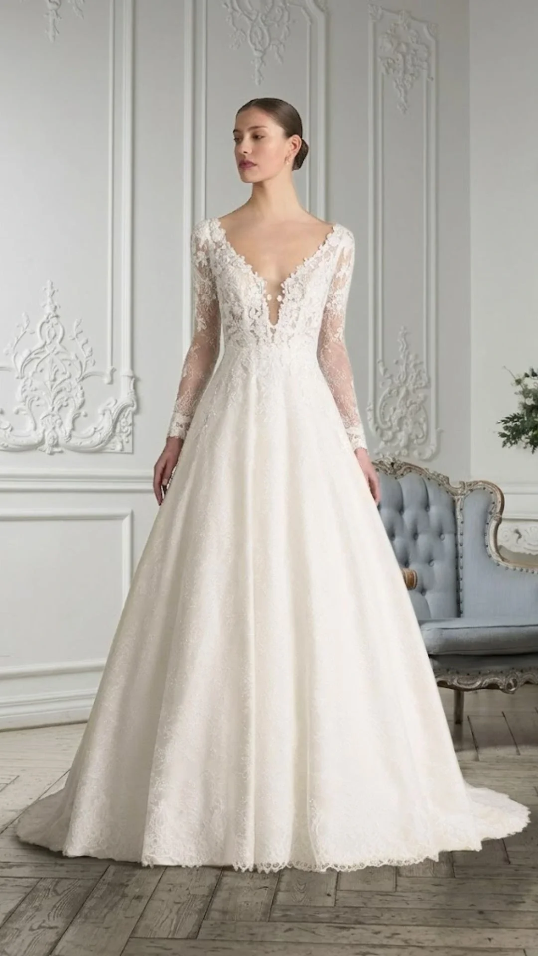 Vestido de noiva com detalhes em renda, decote em V profundo, mangas longas de renda e saia rodada, em um ambiente com molduras brancas no fundo.