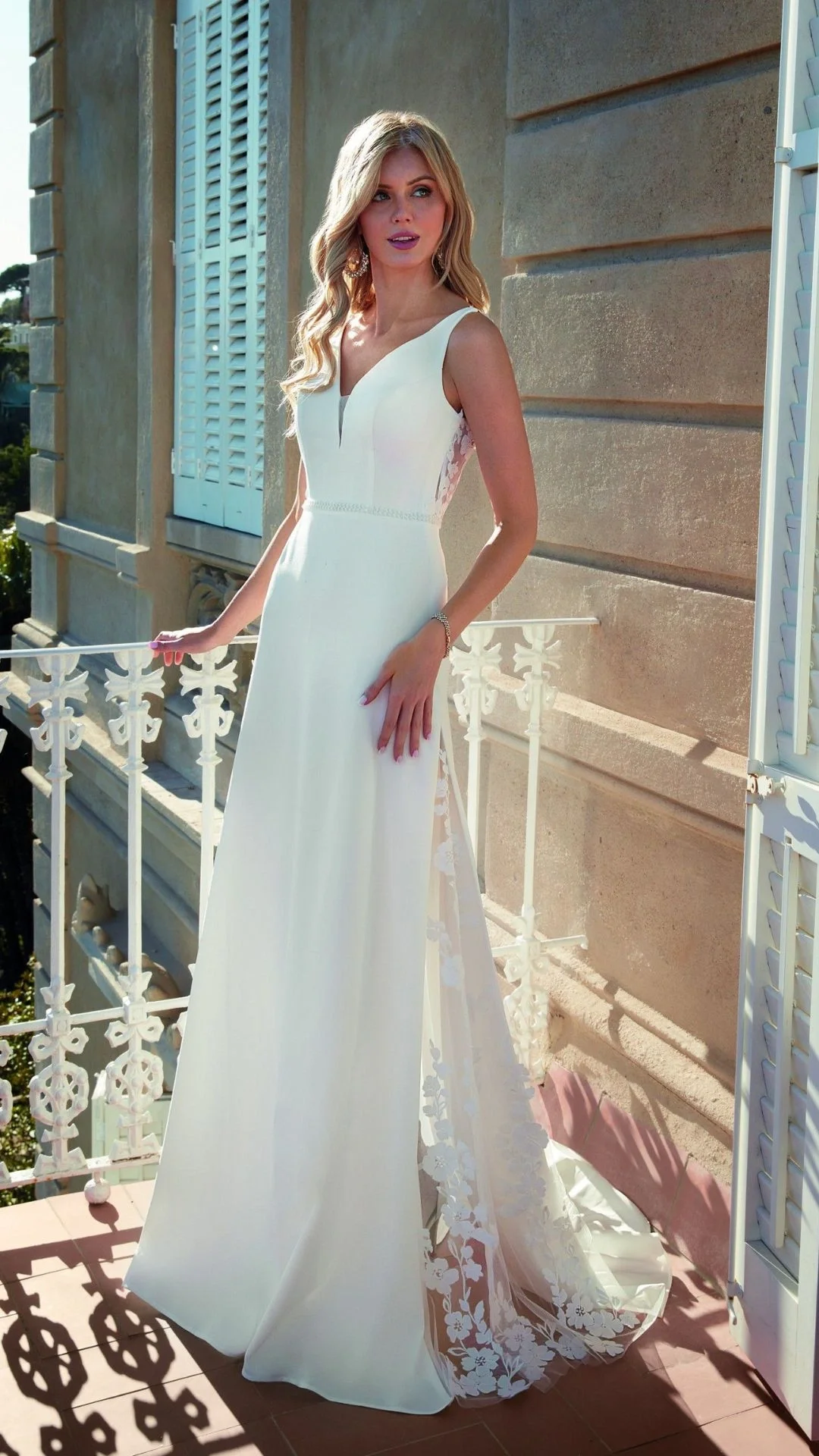 Mulher usando vestido de noiva branco em varanda, com parede de cimento ao fundo e janelas com venezianas brancas.