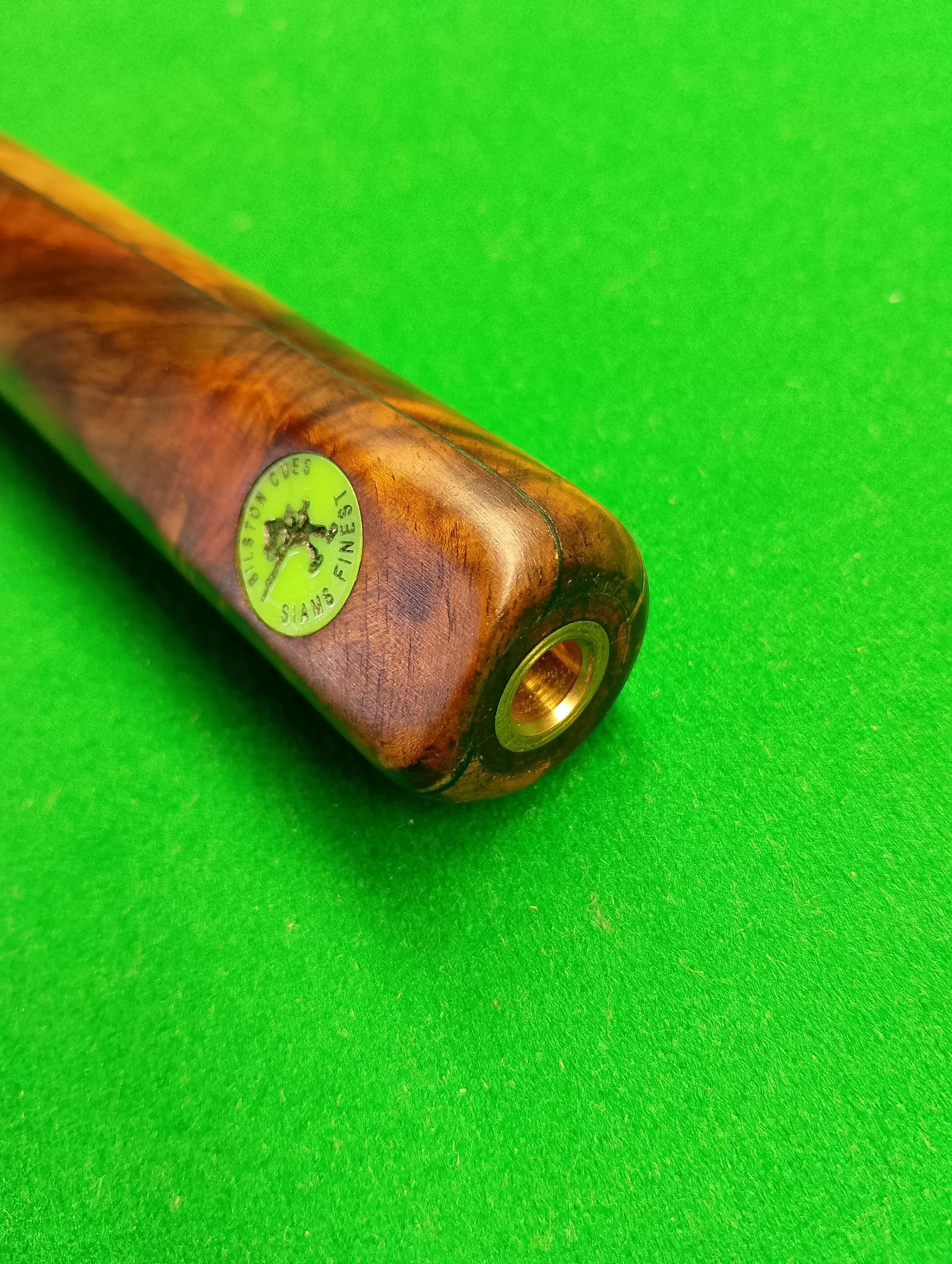 Close-up of a wooden cue stick on a green pool table, with a green sticker reading 'FINEST CUES' and an image of a tree.