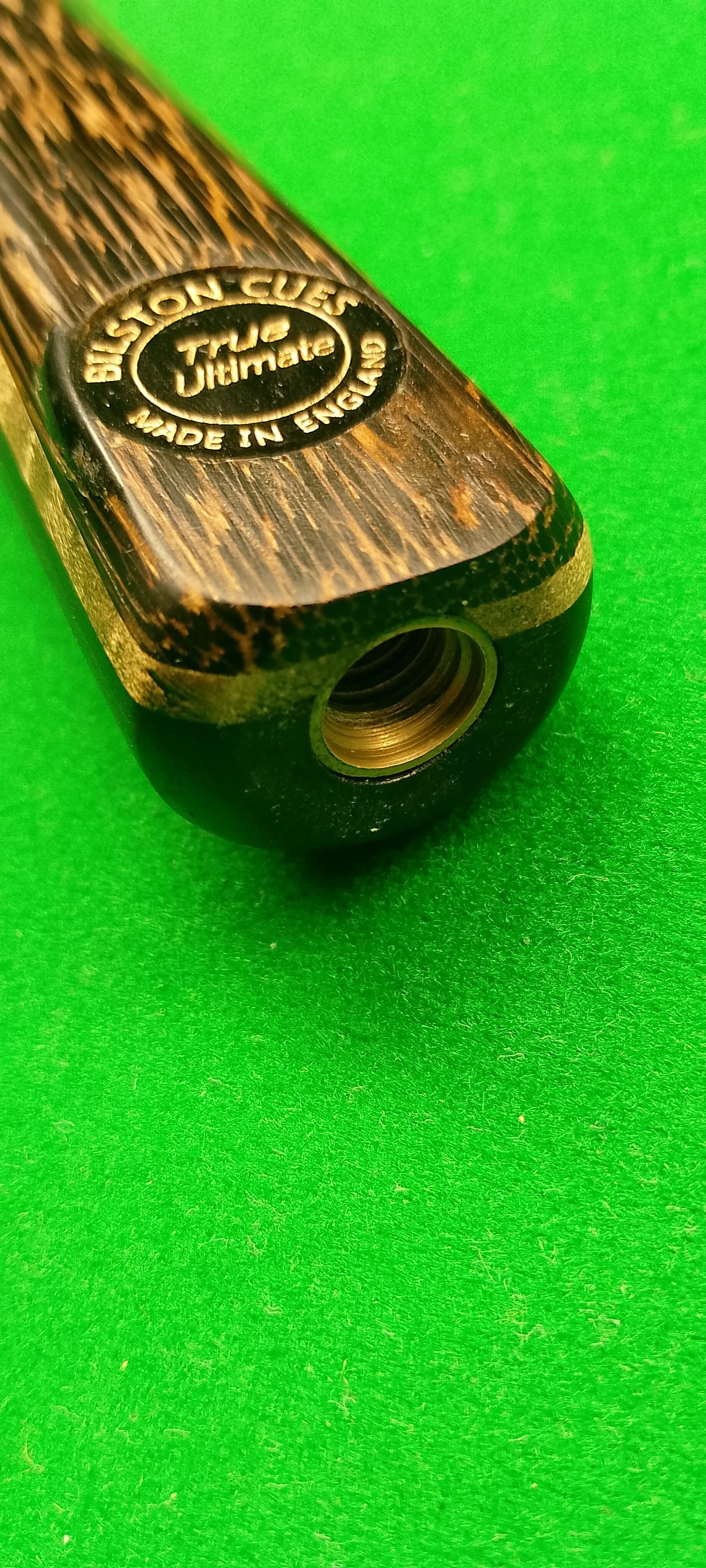 Close-up of a black and wood-textured billiard cue stick with a round emblem that reads 'BILLIARD CLUB, TRUE JIMMY, MADE IN ENGLAND', placed on a bright green surface.