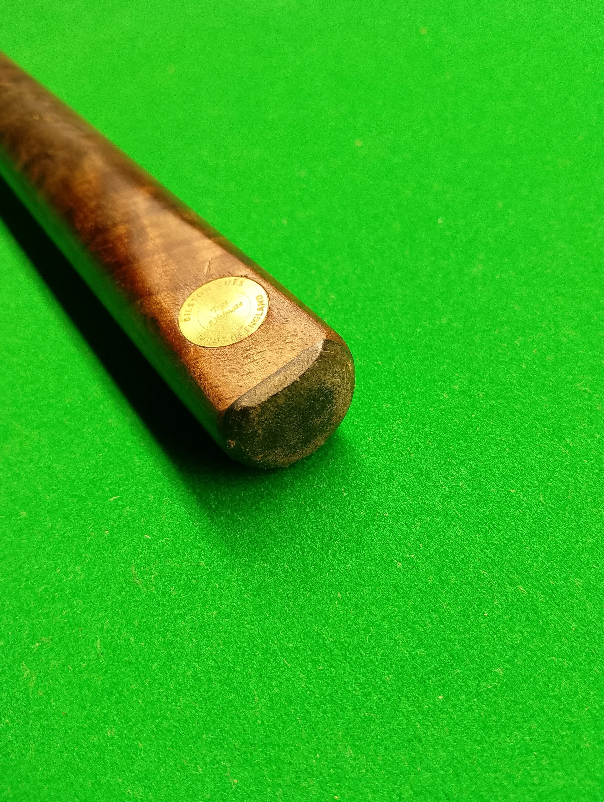 Close-up of a vintage billiard cue resting on a green pool table surface, with a gold sticker indicating it is made in England.