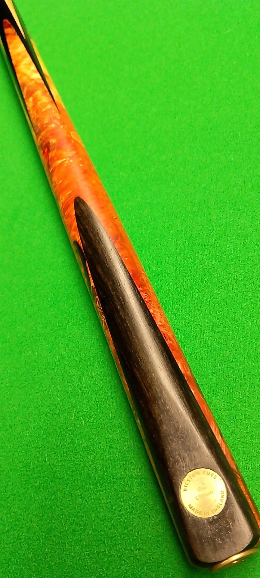 Close-up of a snooker cue with a wooden finish and a gold sticker, placed on a bright green snooker table.