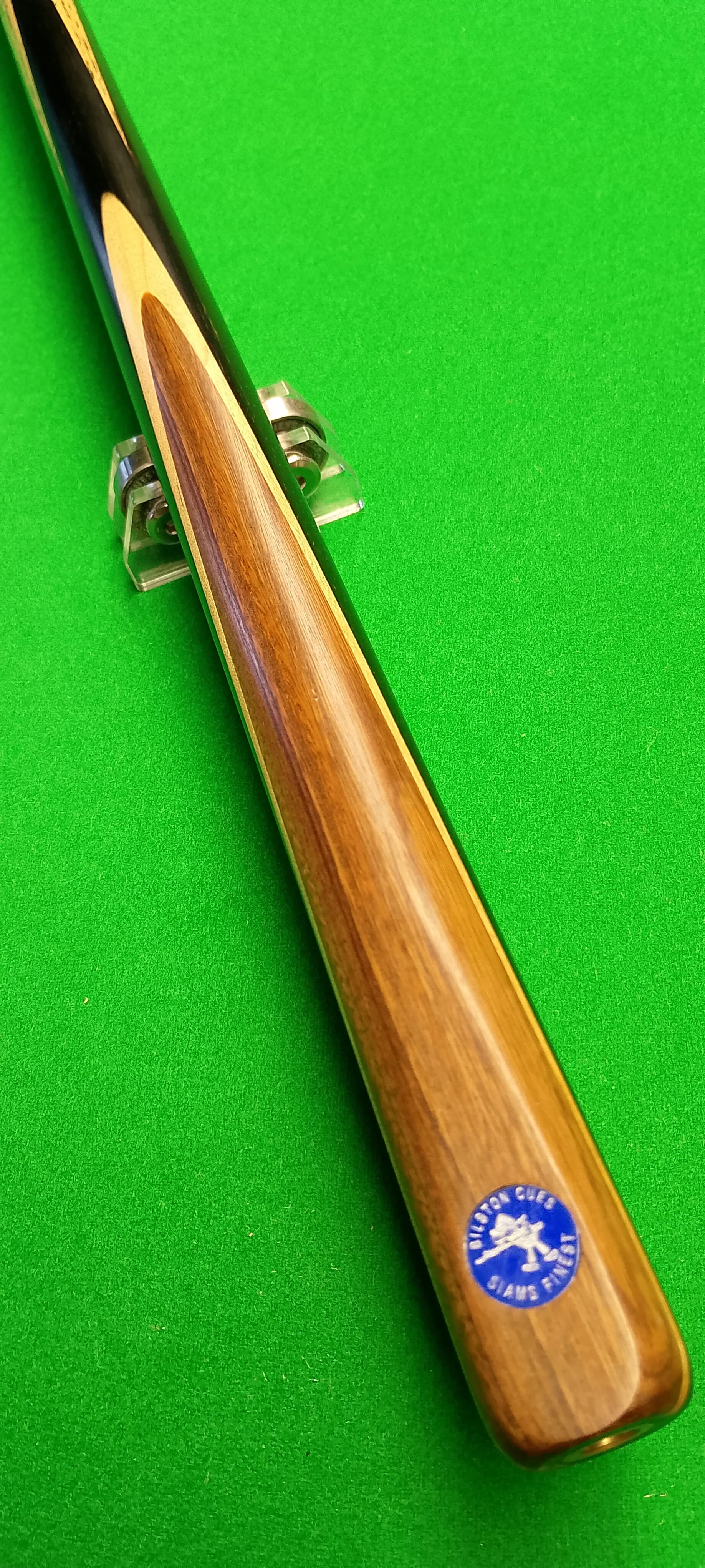 A wooden cue stick for billiards or pool resting on a green pool table surface with a sticker near the butt end.