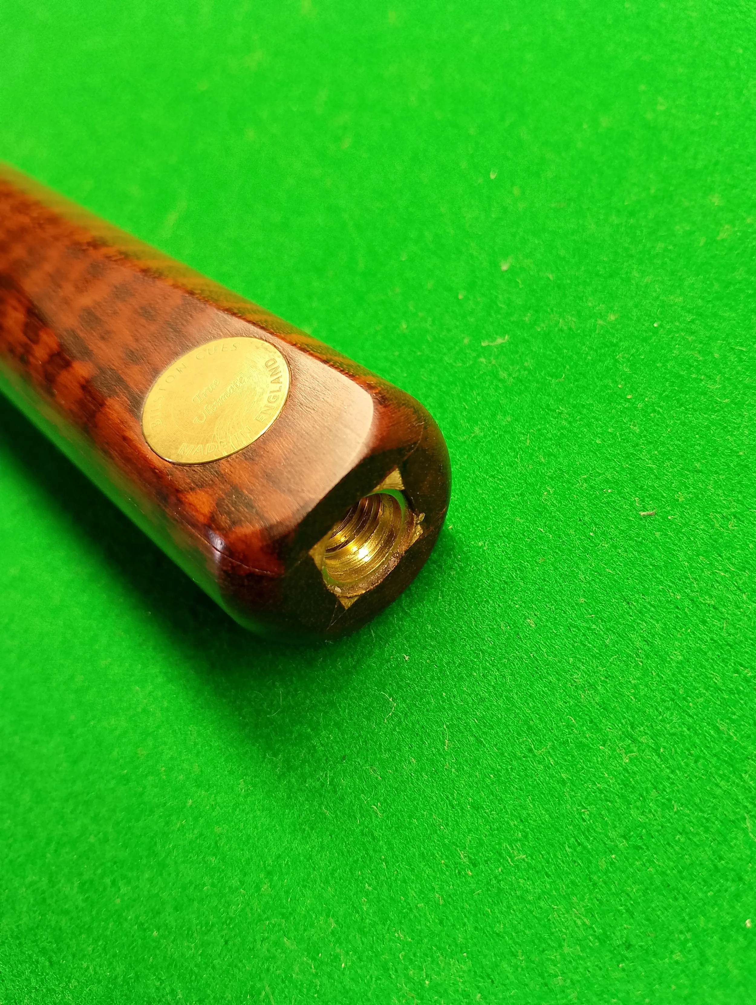Close-up of a wooden exoskeleton pool cue with a metal screw thread at the end and a gold-colored sticker that reads 'DIRECTION USE True Durtness MADE IN ENGLAND' on a green felt surface.