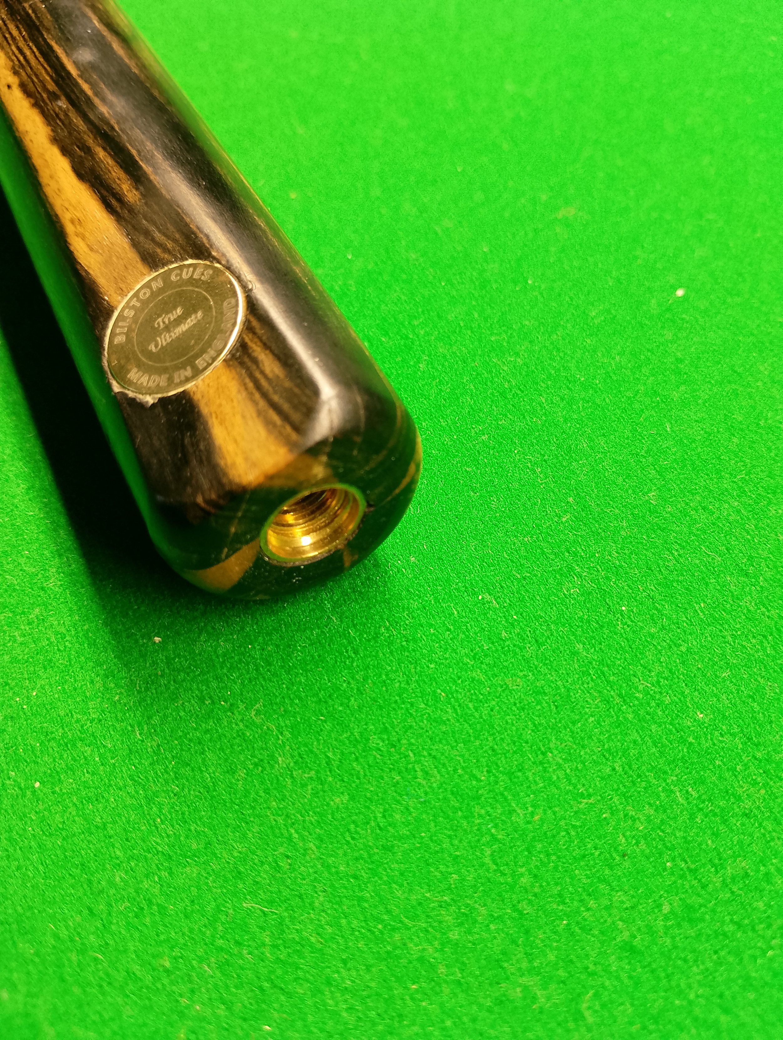 Close-up of a billiard cue stick with a metal sticker, resting on a green pool table felt, showing the cue's bottom with a threaded screw hole.
