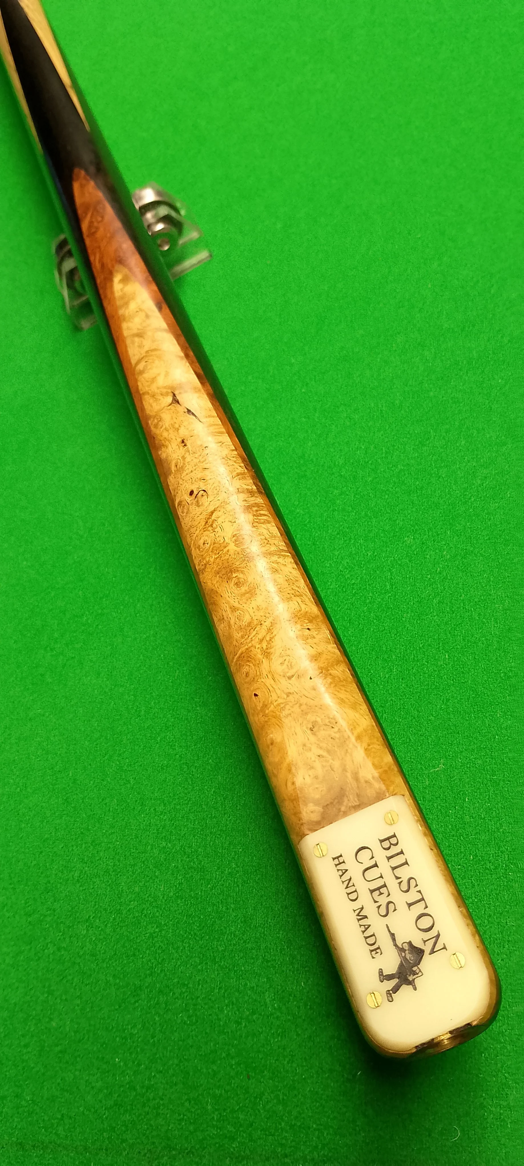 Close-up of a billiard cue with a wood grain handle on a green pool table felt. The cue has a metal joint and a white joint cap with the logo and text 'BUSTON CUES HAND MADE'.