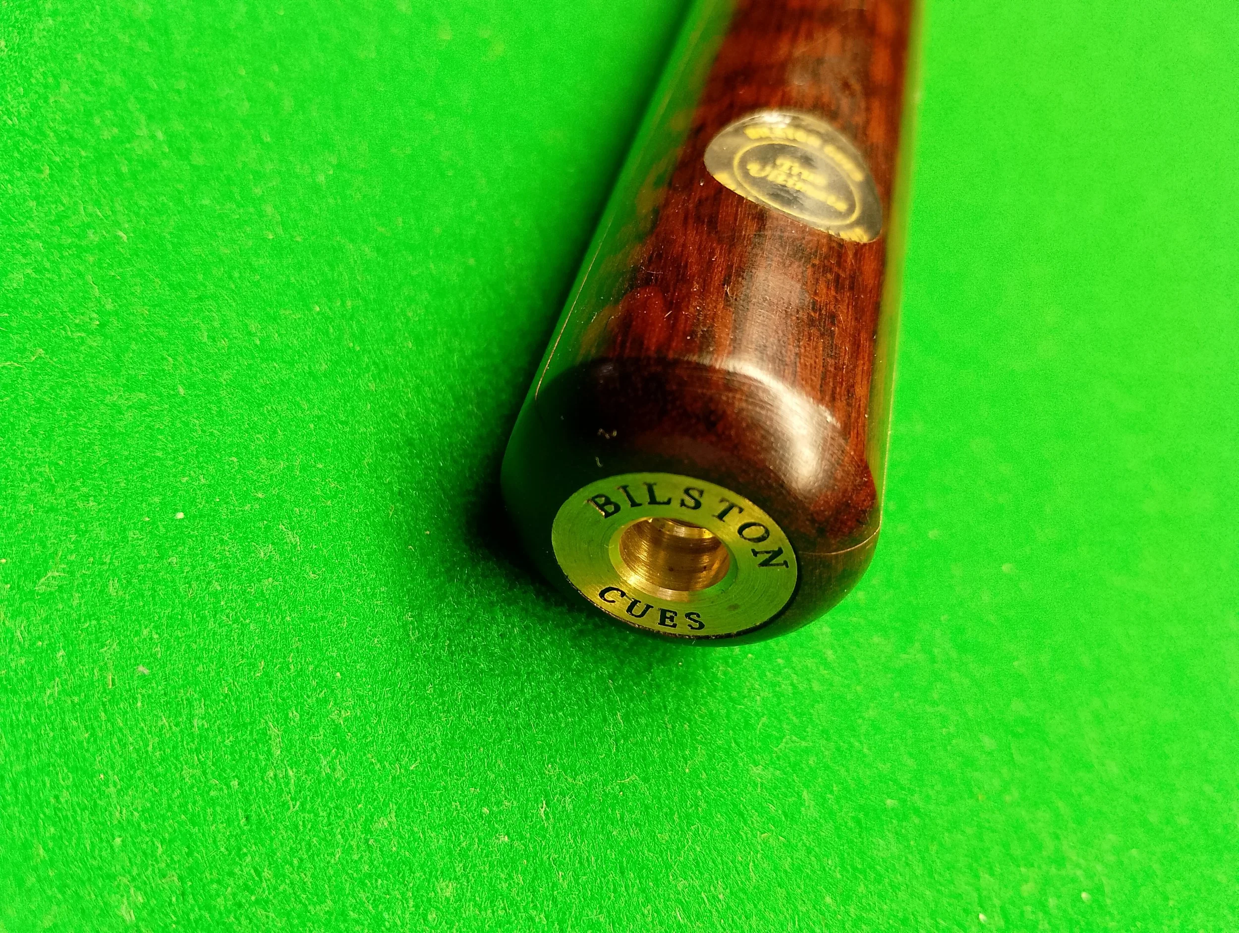 Close-up of a billiard cue end with metal fitting labeled 'BILSTON CUES' on a green billiard table.