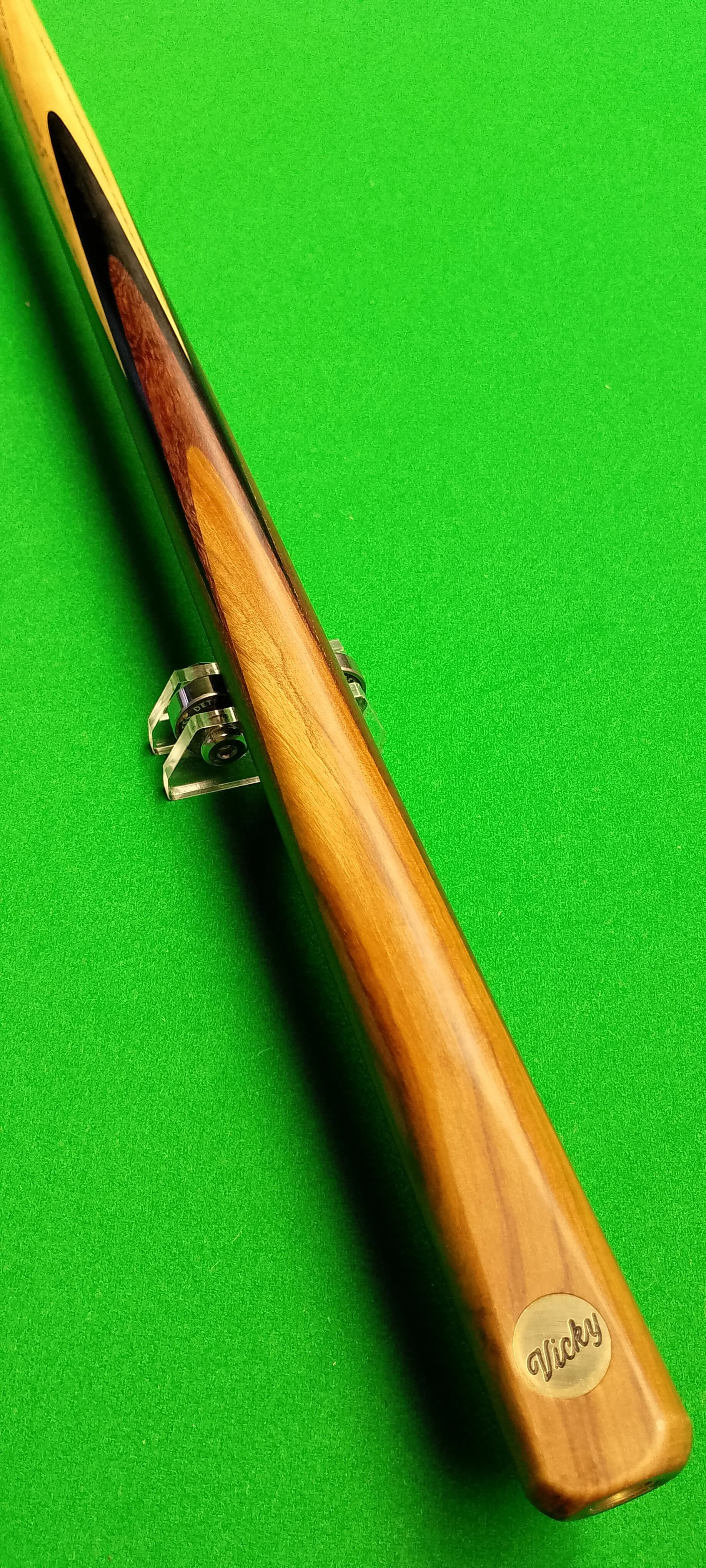 A custom wooden rifle with a natural wood finish and a metal medallion engraved with the name 'Vicky' on the stock, displayed on a green background.