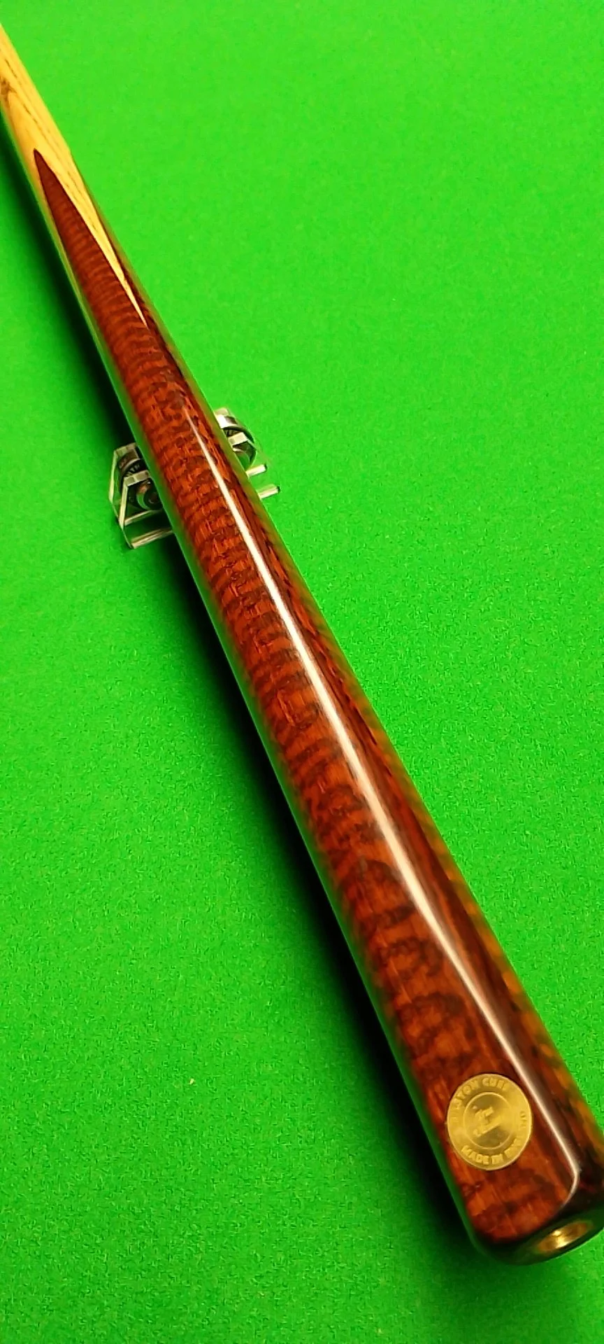 A wooden cue stick with a shiny finish and a small round gold sticker, resting on a green surface.