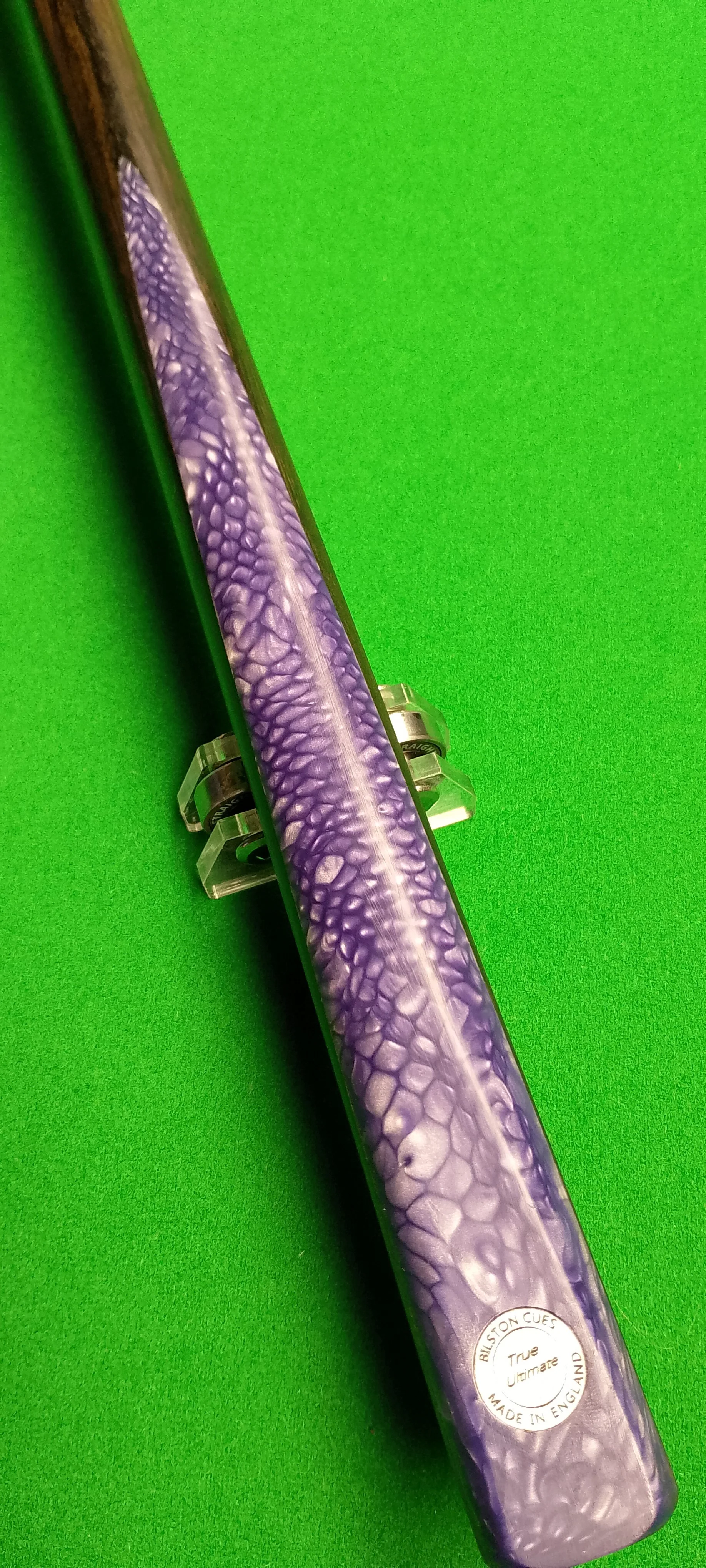 Close-up of a cue stick with a purple and white pattern, placed on a green surface, with a sticker that says 'BUSTON CUES, True Ultimate, Made in England'.