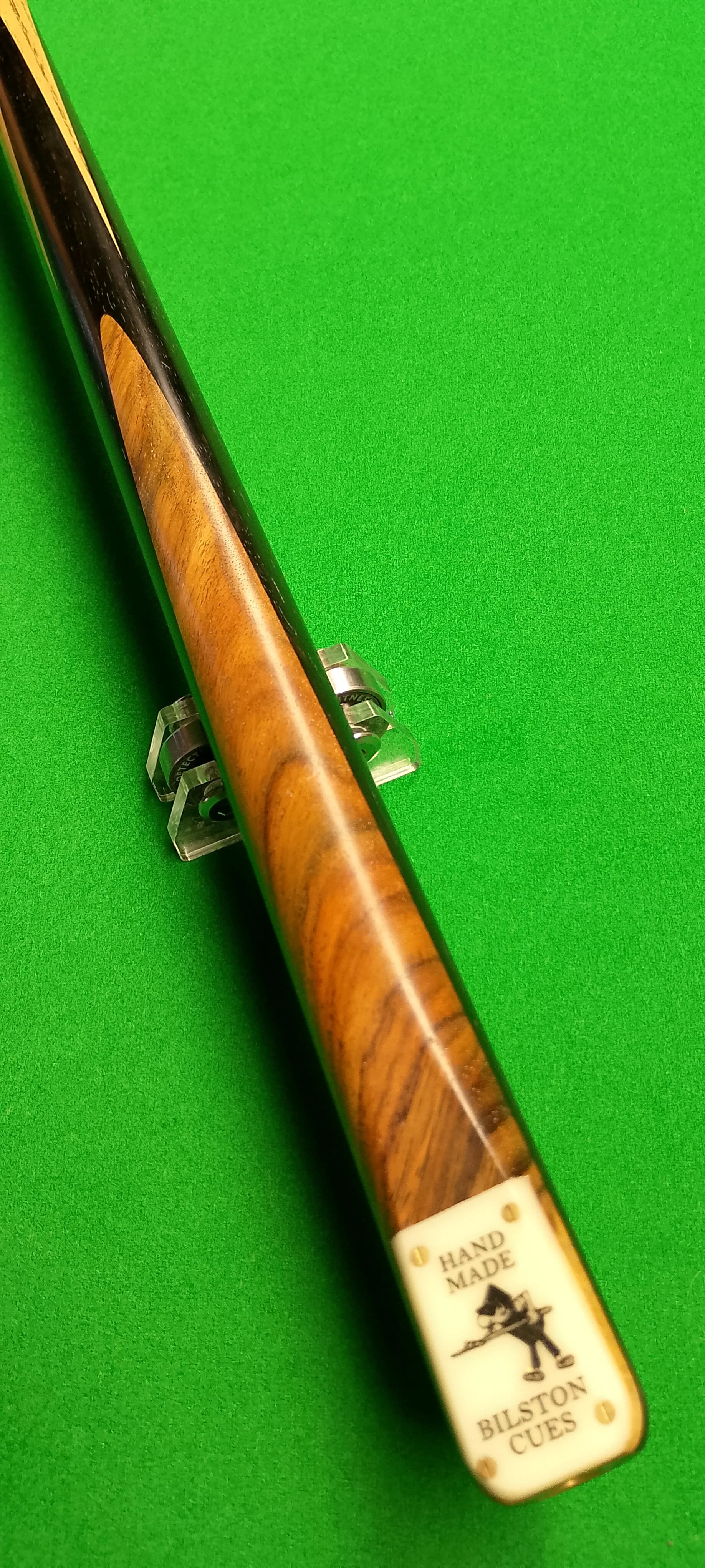 A professional pool cue with a wooden finish and a white end cap that has a cartoon character holding a cue stick, along with text that reads 'Hand Made Bilston Cues', placed on a green pool table.