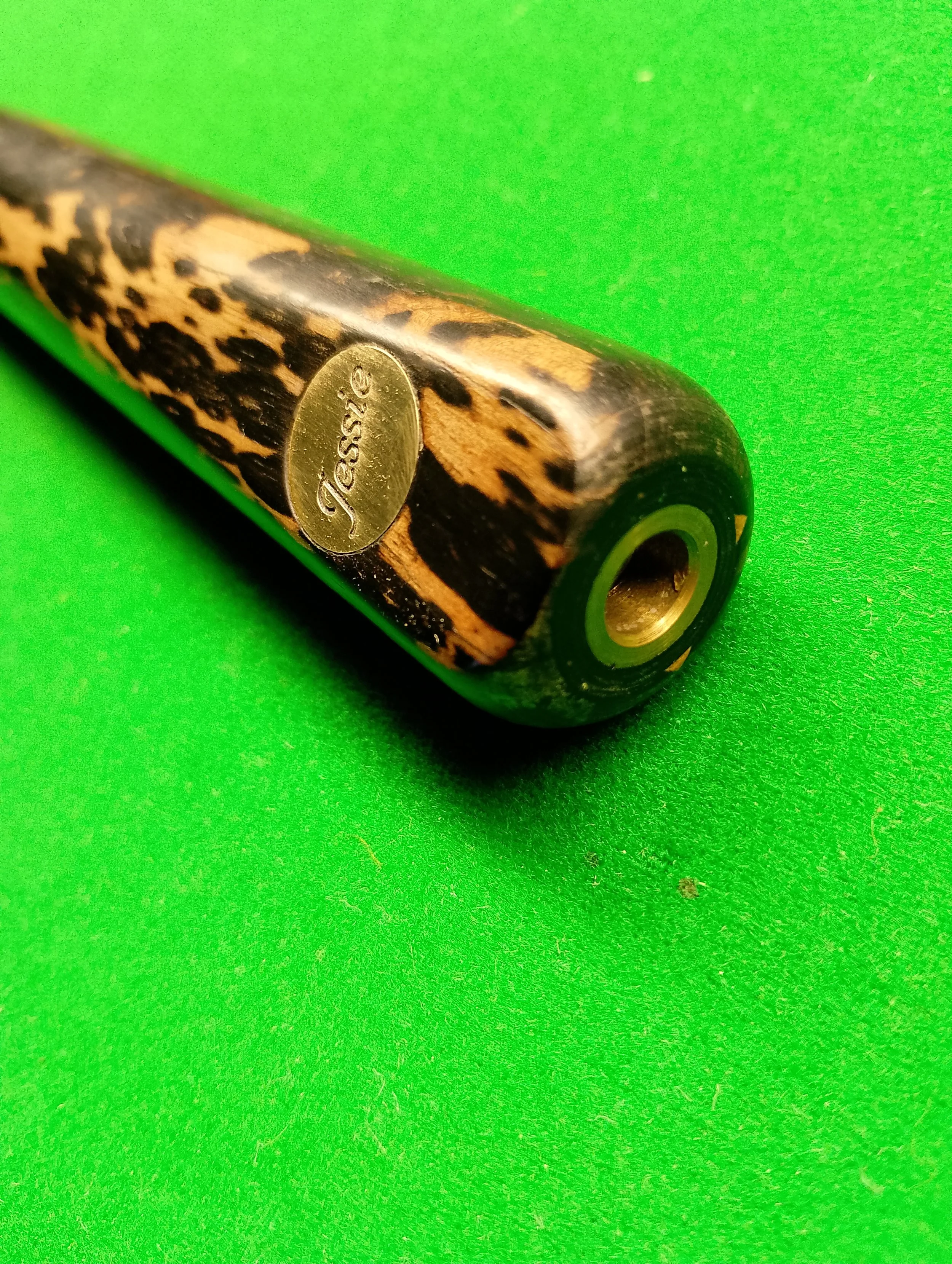 Close-up of a pool cue handle with a leopard print design and a gold-colored "Jesse's" oval emblem, on a green billiard table felt.