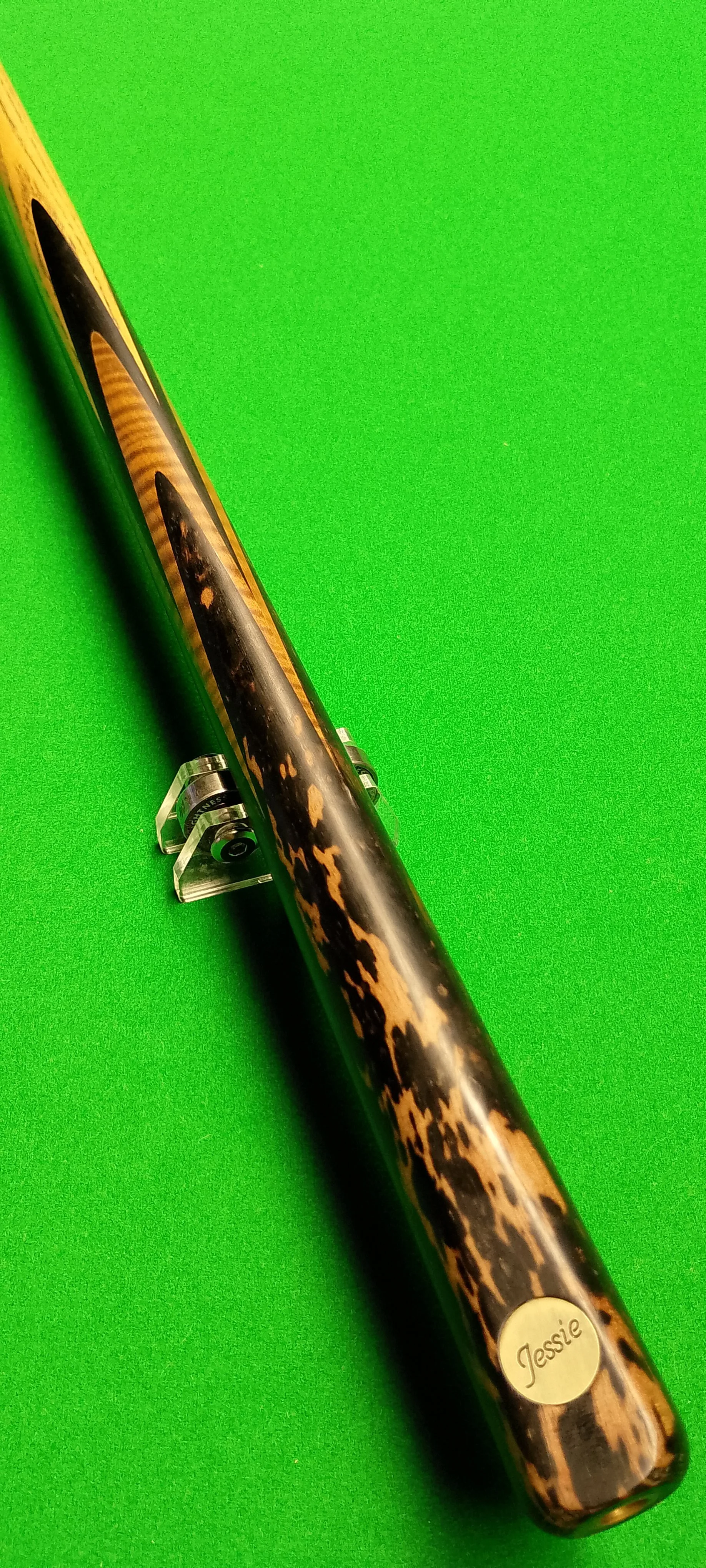 Close-up of a pool cue with a tortoise shell pattern, labeled with the name "Jessie," resting on a green pool table.