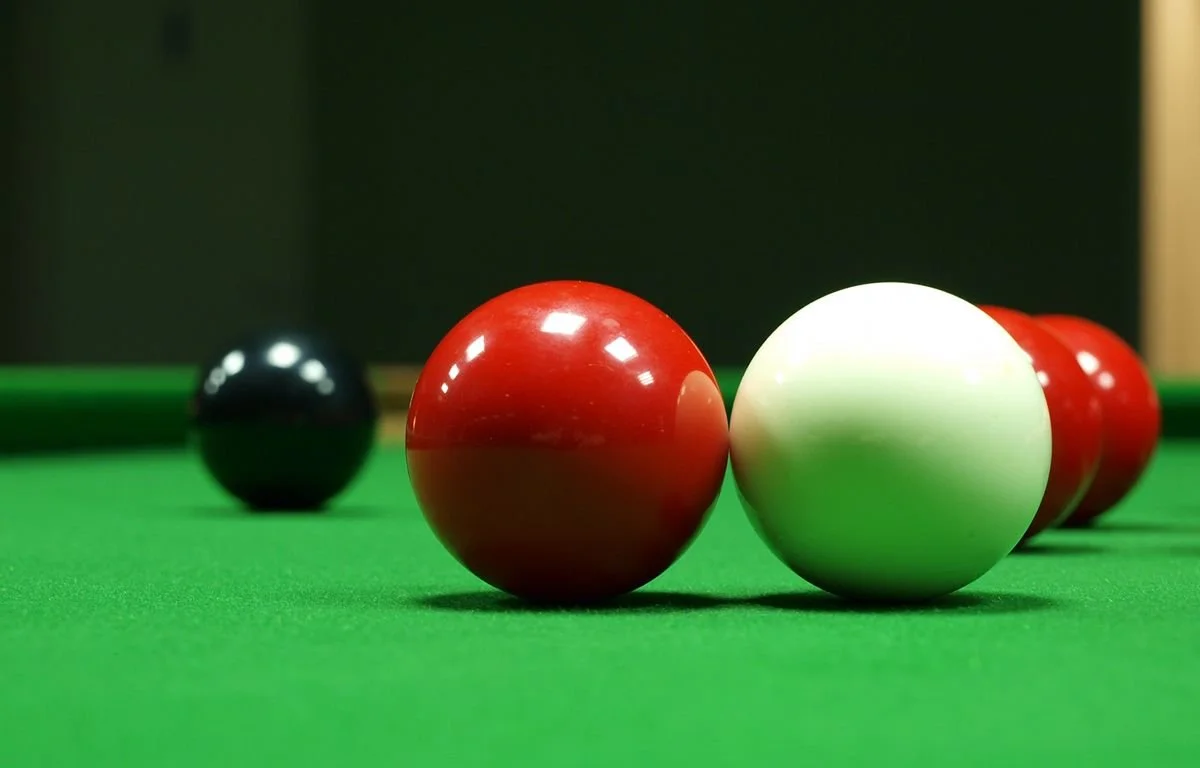 Close-up of snooker or pool balls on a green felt table, with a black ball in the background and a red, white, and more red balls nearby.