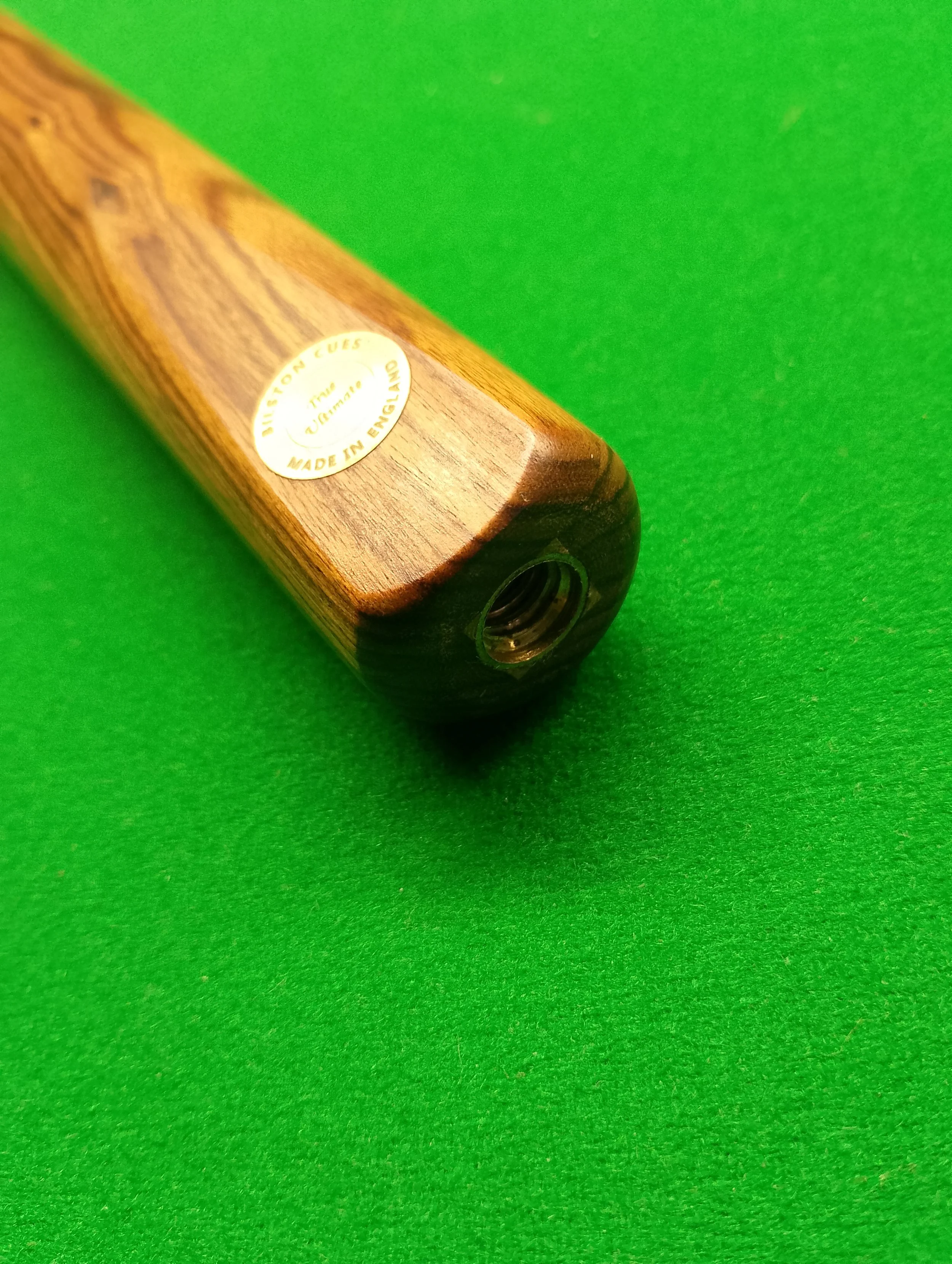 Close-up of a wooden snooker cue end with a circular sticker labeled 'Billesdon Cues, The Ulimate, Made in England,' on a green felt surface.