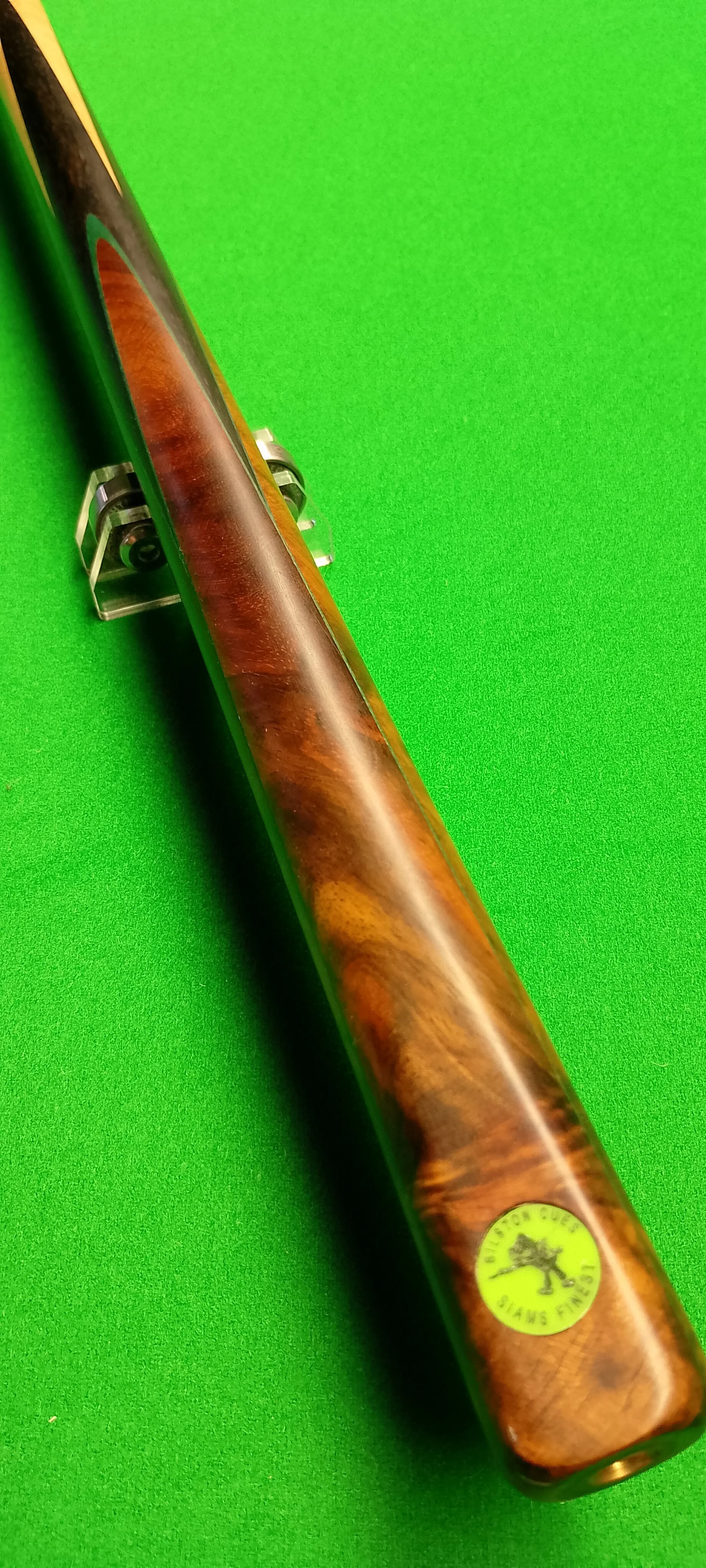 A wooden cue stick on a green pool table with a sticker at the bottom end that reads 'Player's Cue by Sure Shot' with a lion illustration.