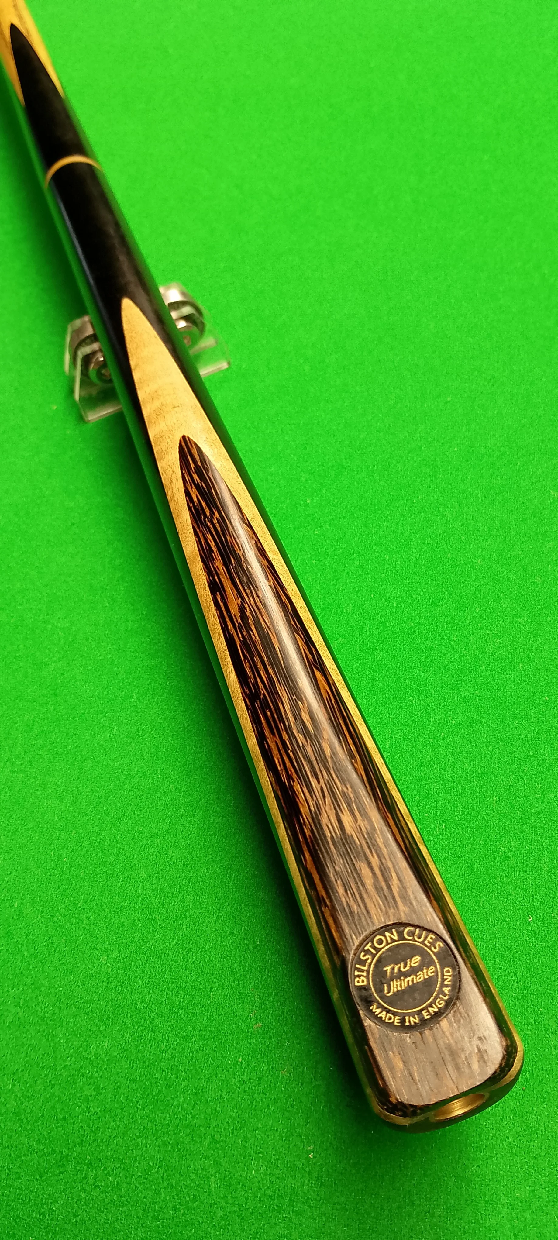 A cue stick with a black and gold color scheme, resting on a green billiard or pool table felt. The cue has a decorative wooden inlay near the tip and a circular label near the butt end that reads "Billston Cues, True Ultimate, Made in England."
