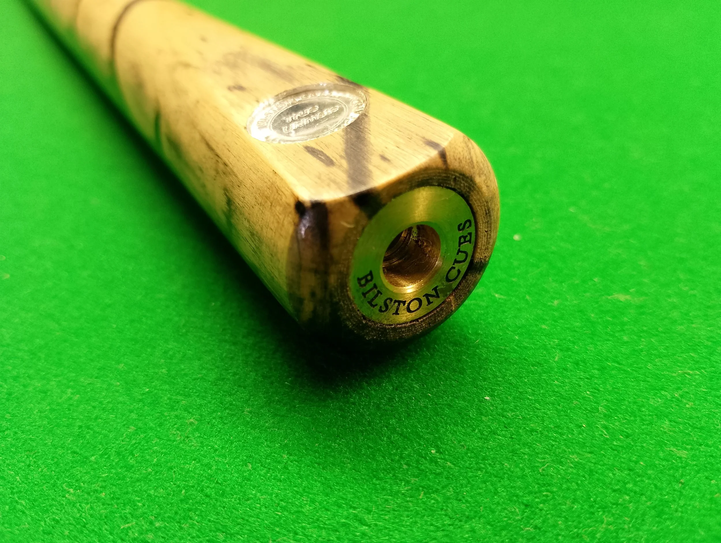 A close-up of a brass pool cue stick with a round determined for cue tip and the inscriptions 'BOSTON CUES' around the inner edge, lying on a green felt surface.