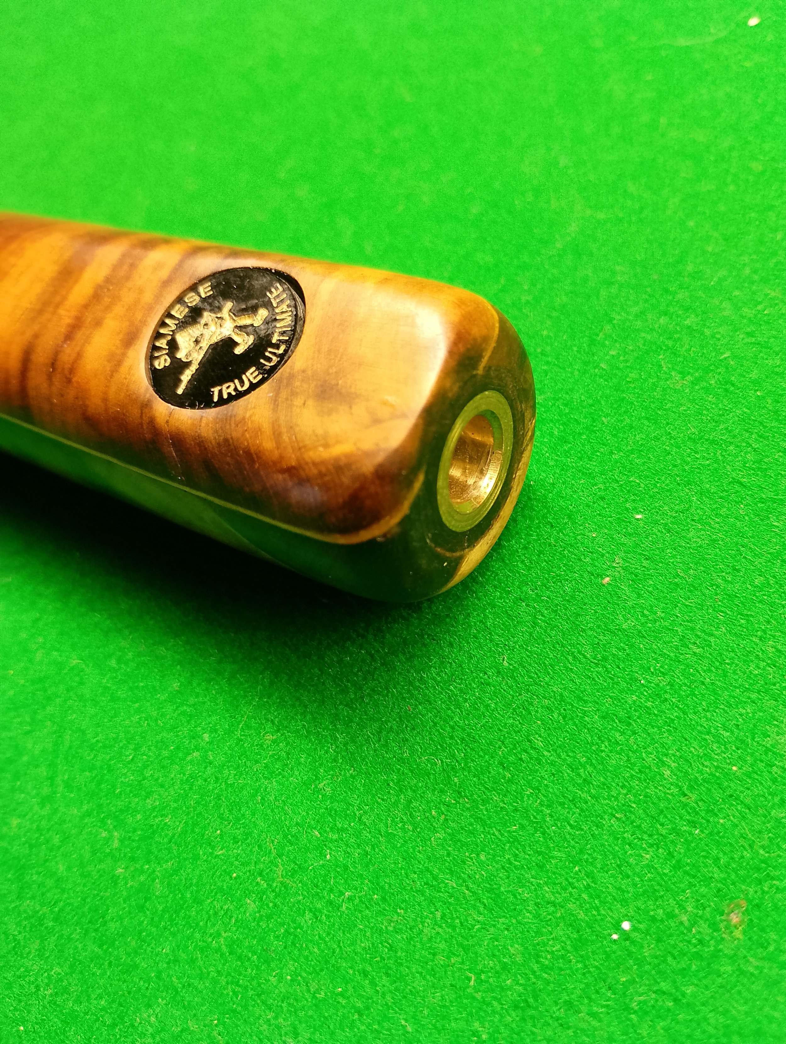 Close-up of a wooden cue stick with a black circular logo that says 'Shaneise True Uline' with a lion logo, resting on a green pool table.