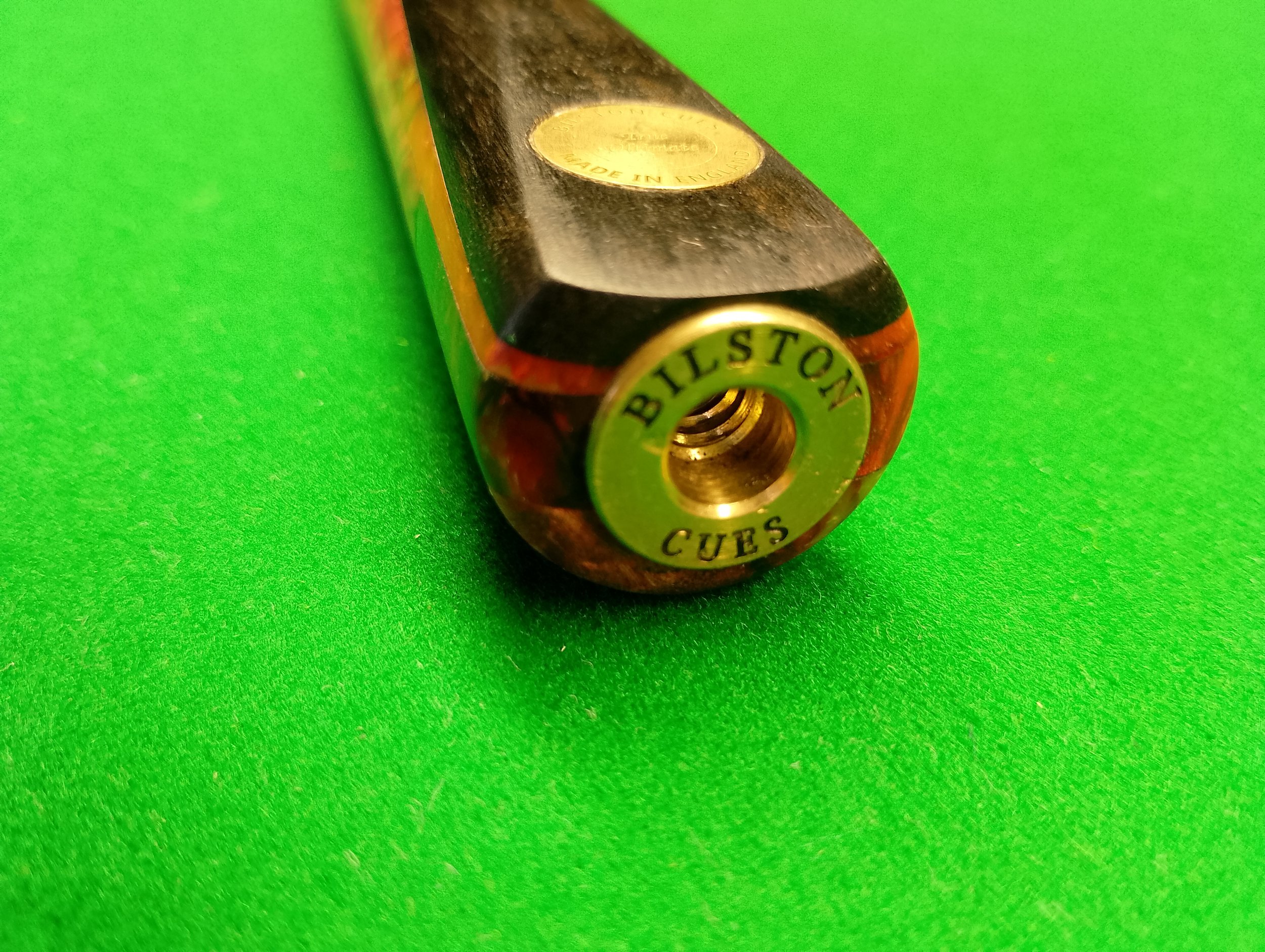 Close-up of a billiard cue stick end with a metal joint labeled 'BILSTOI CUES' on a green felt billiard table.