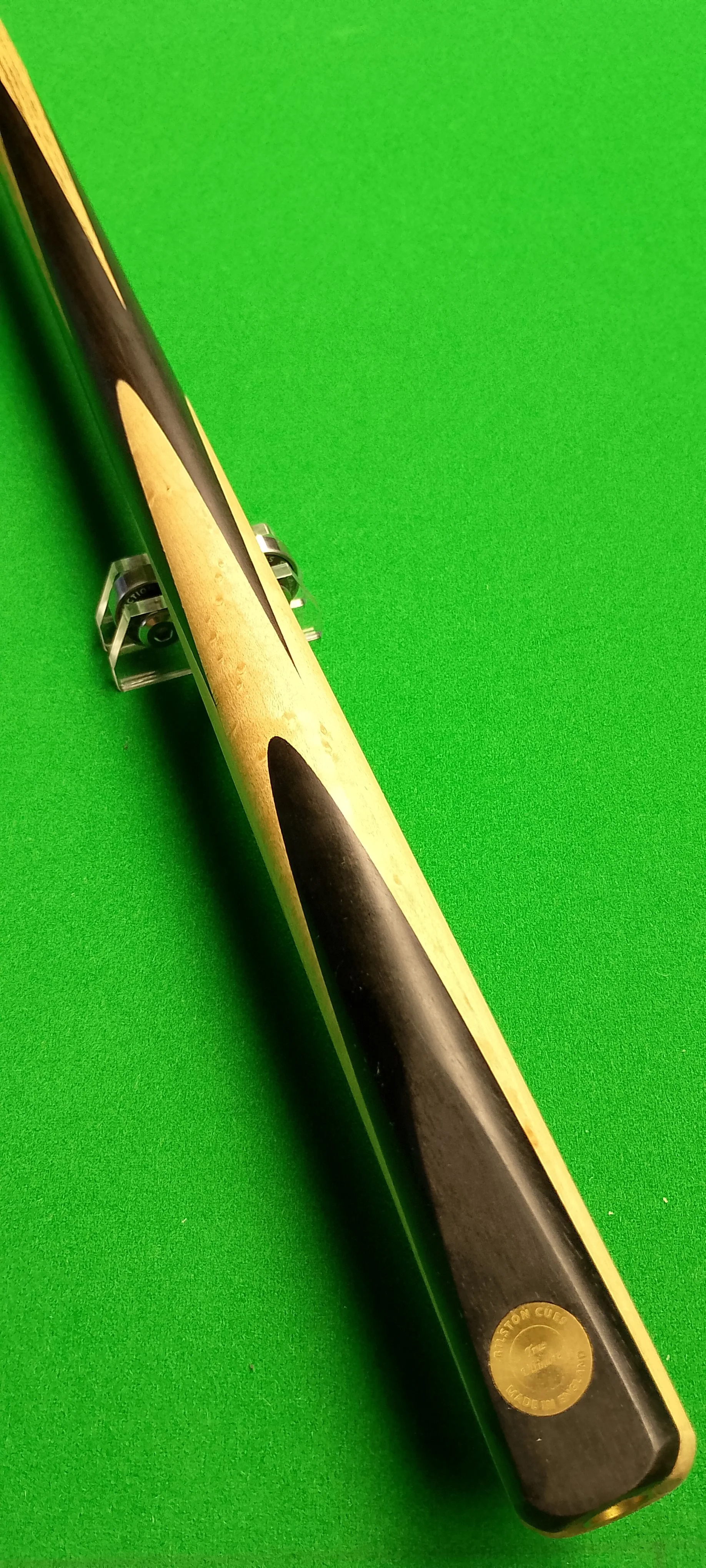 A pool cue is positioned on a green pool table. The cue has a black tip and a light-colored shaft. The butt features a black and gold design with the brand name "St. Louis" visible.