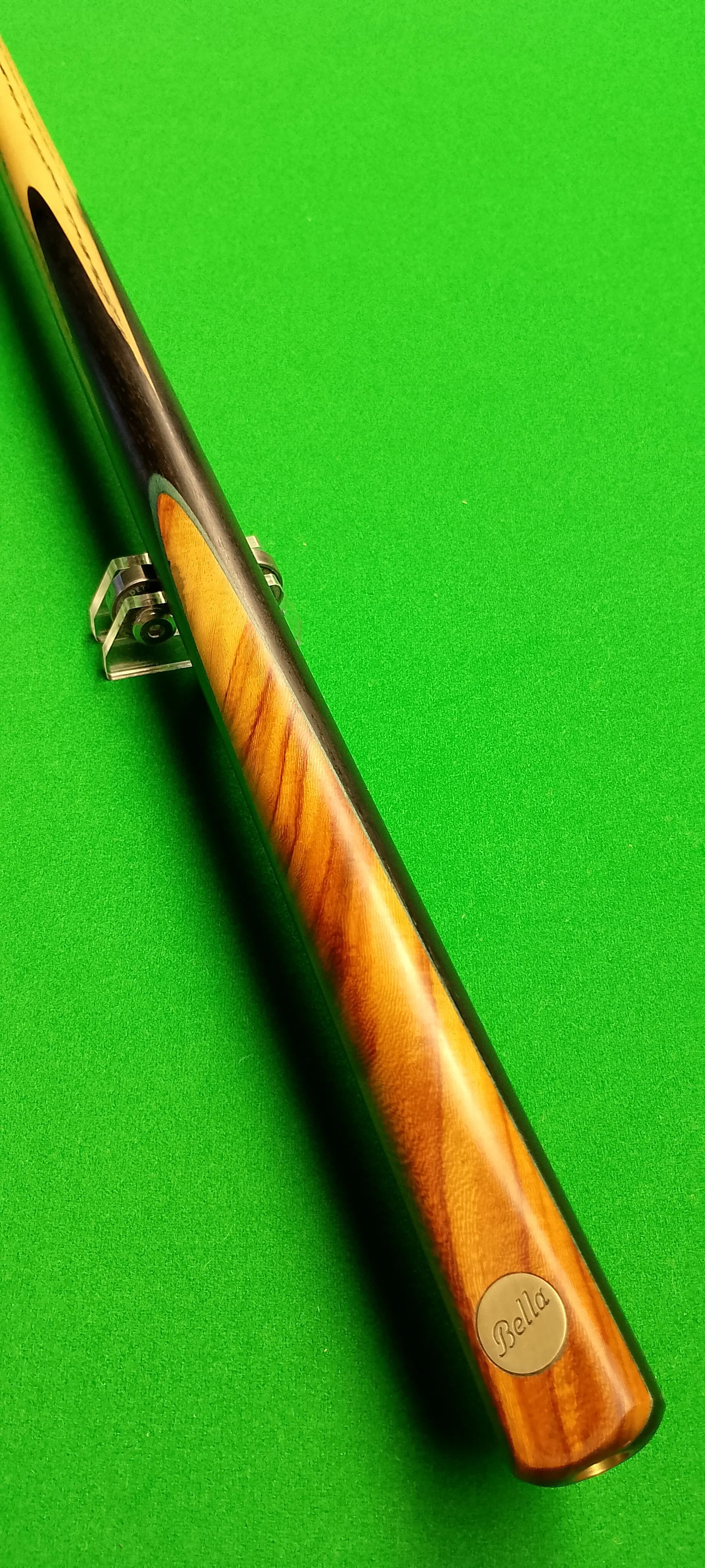 A pool cue with a wooden handle and a metal tie engraved with the name 'Bella', resting on a green pool table.