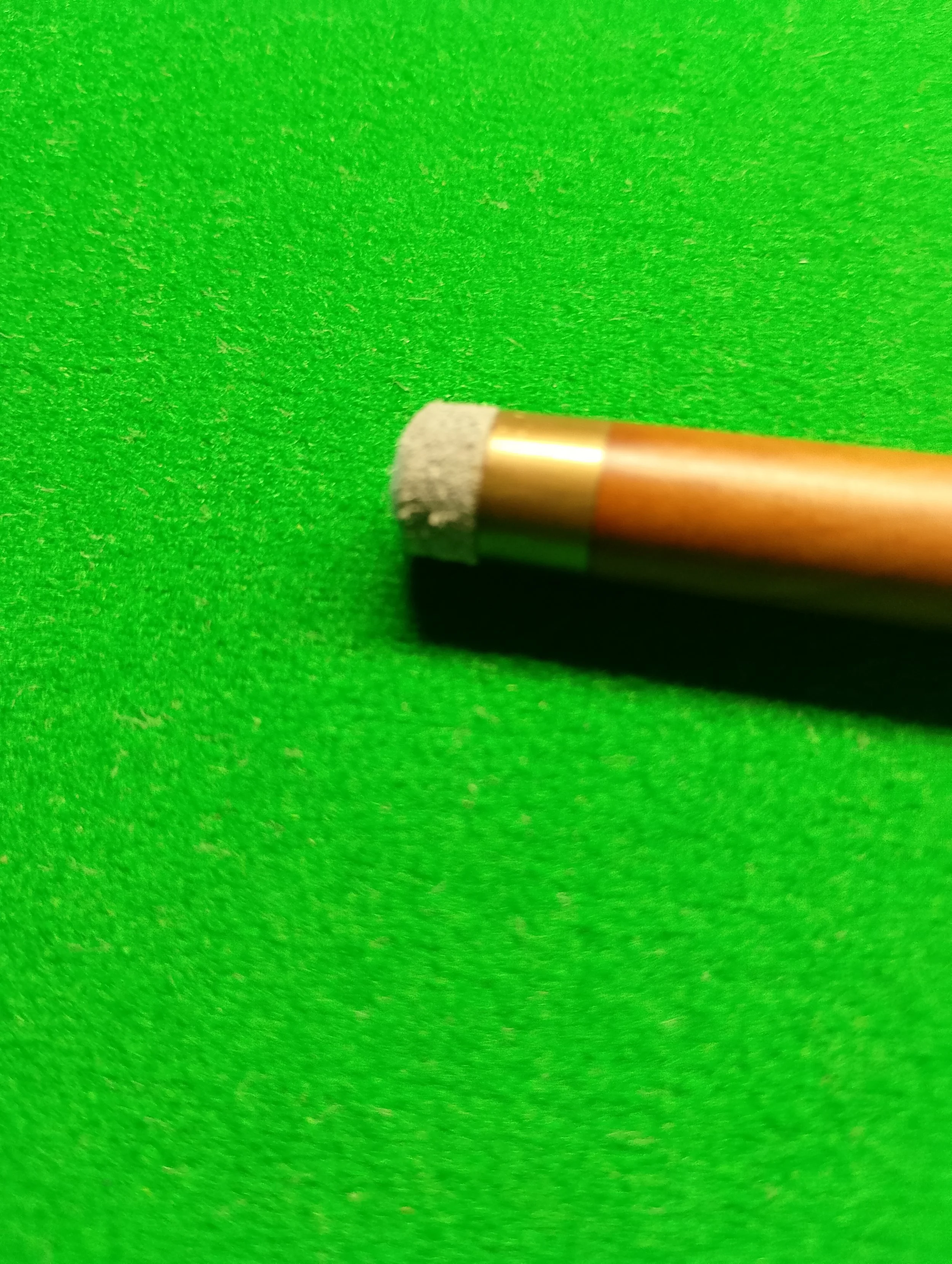 Close-up of a wooden cue stick with a gray chalk tip on a green felt pool table.