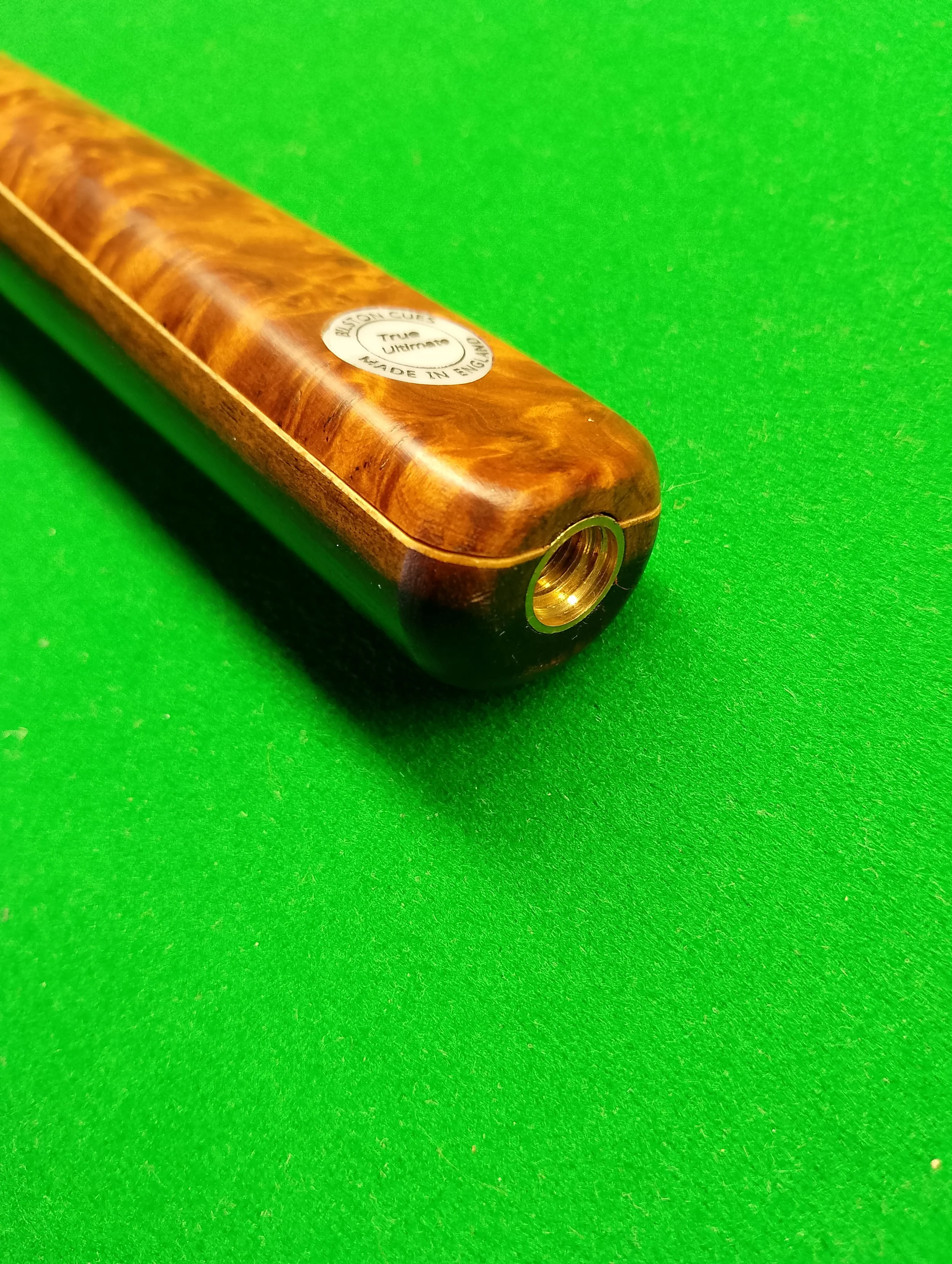 Close-up of a wooden pool cue on a green felt table, showing the tip and a sticker that says 'BASSON CUES, TOP ULTIMATE, MADE IN ENGLAND'.