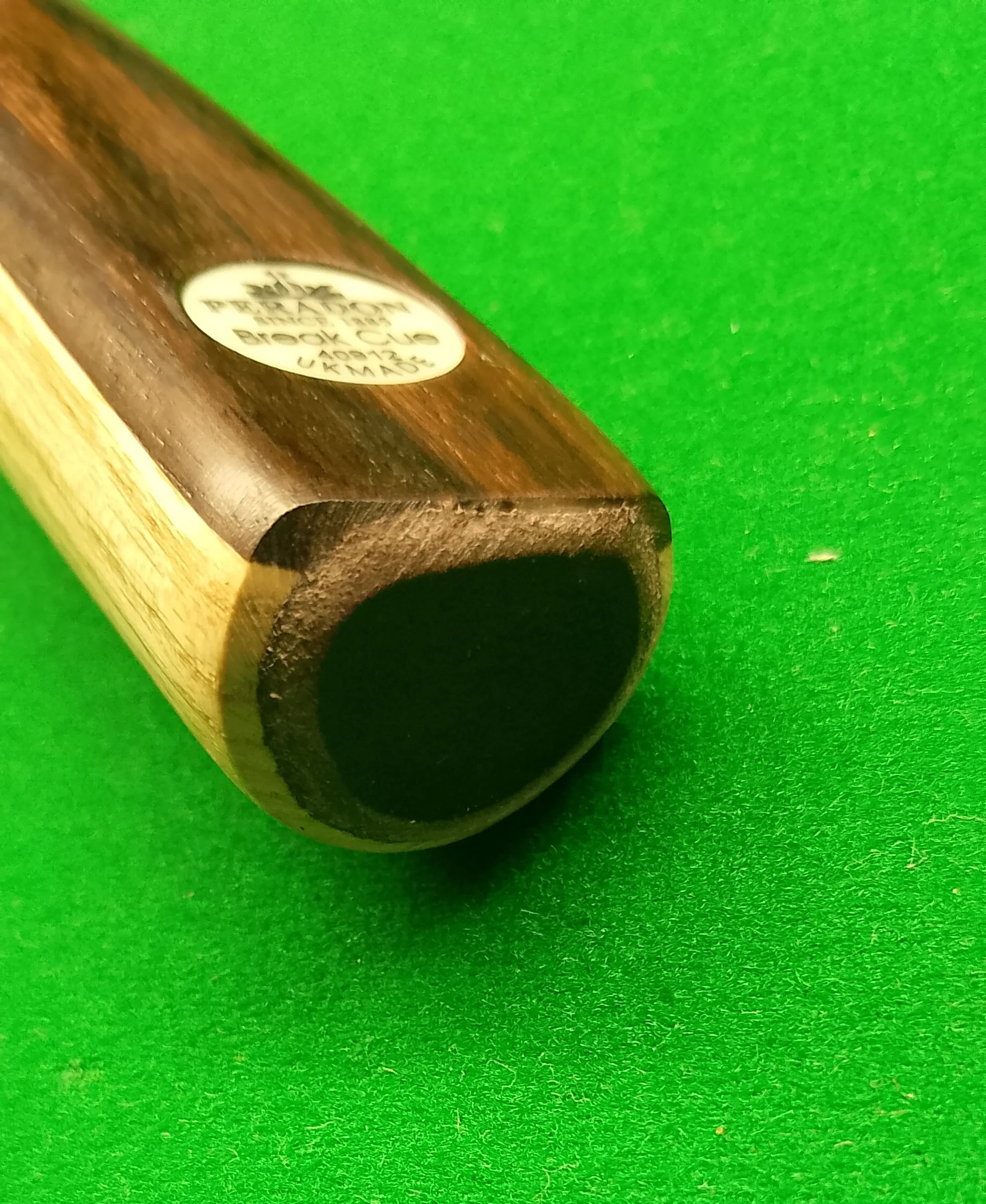 Close-up of the end of a wooden pool cue stick on a green felt surface, with a white sticker containing black text.