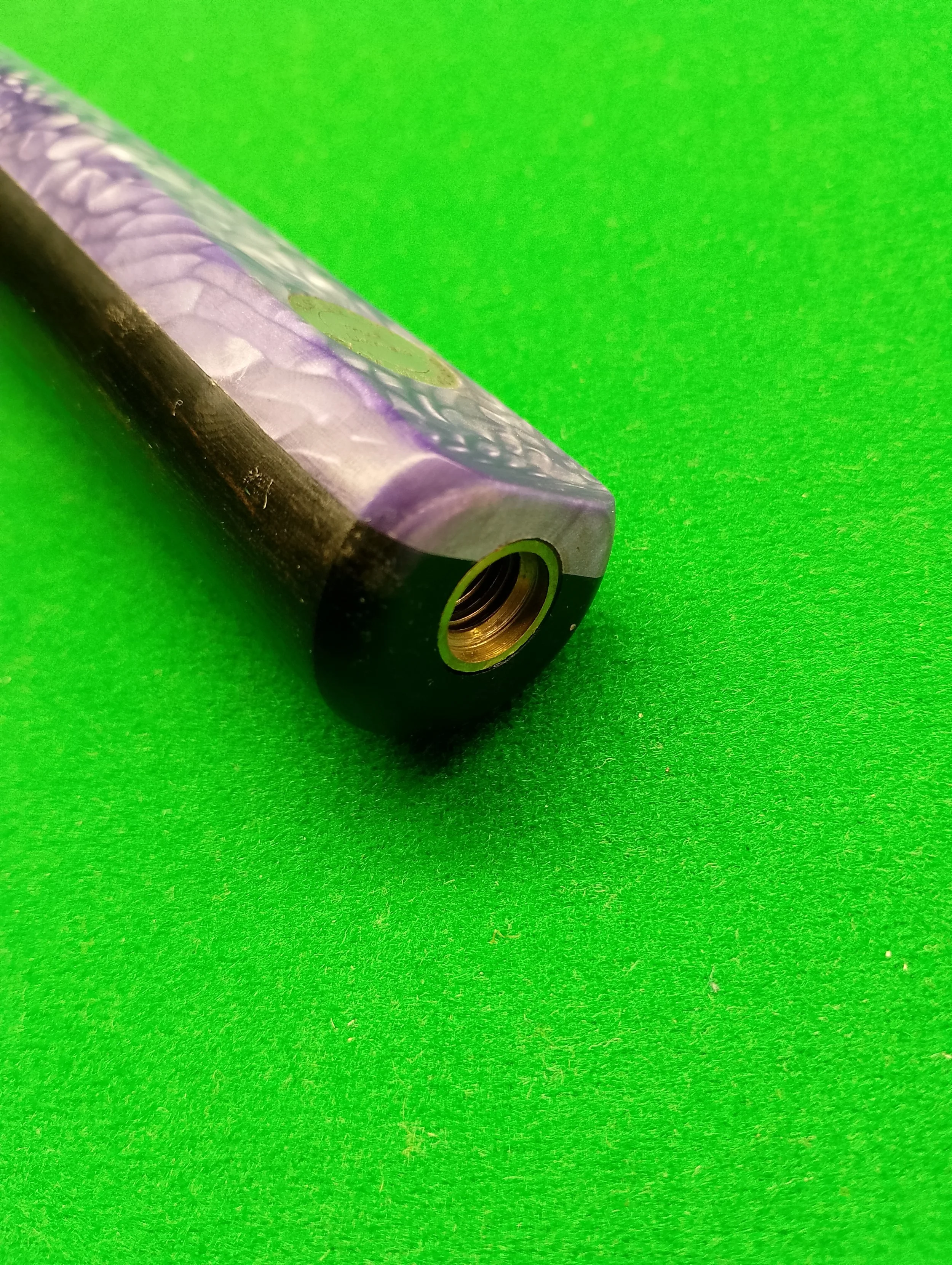 Close-up of a purple and black vape cartridge with a brass threaded connector, lying on a green surface.