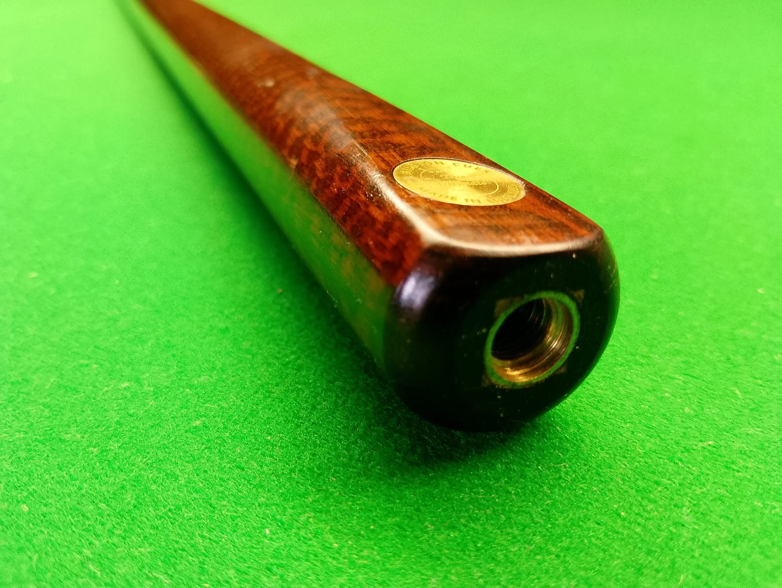 Close-up of a wooden billiards cue stick lying diagonally on a green pool table, with the tapered end facing the camera.