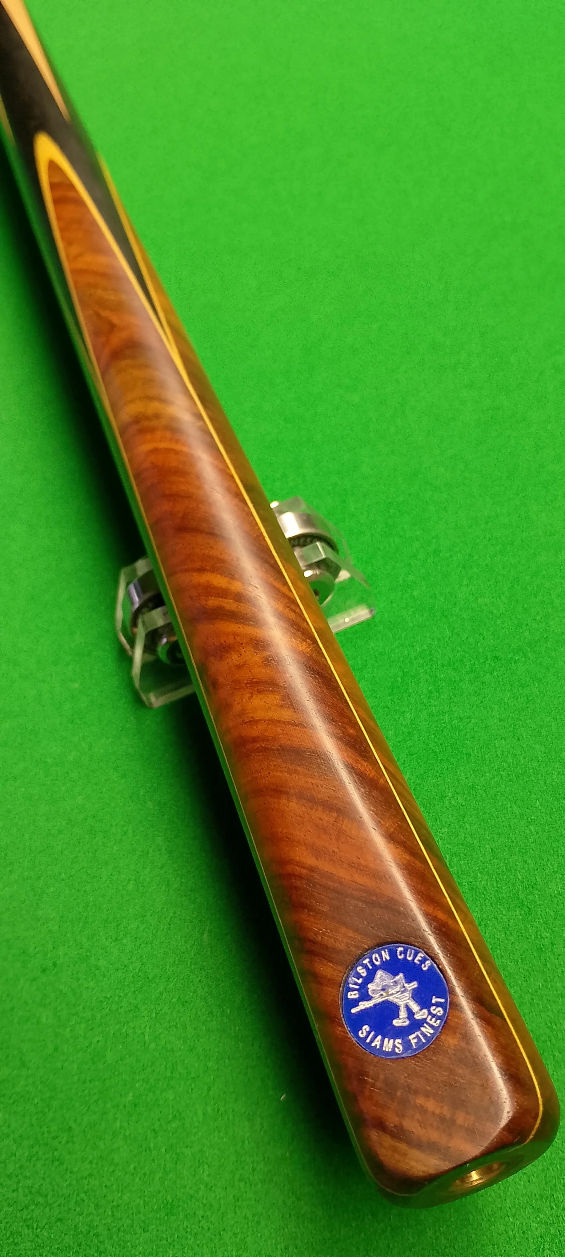 Close-up of a wooden cue stick with a blue sticker that reads 'Briston Cues, Slam's Finest' on a green pool table surface.