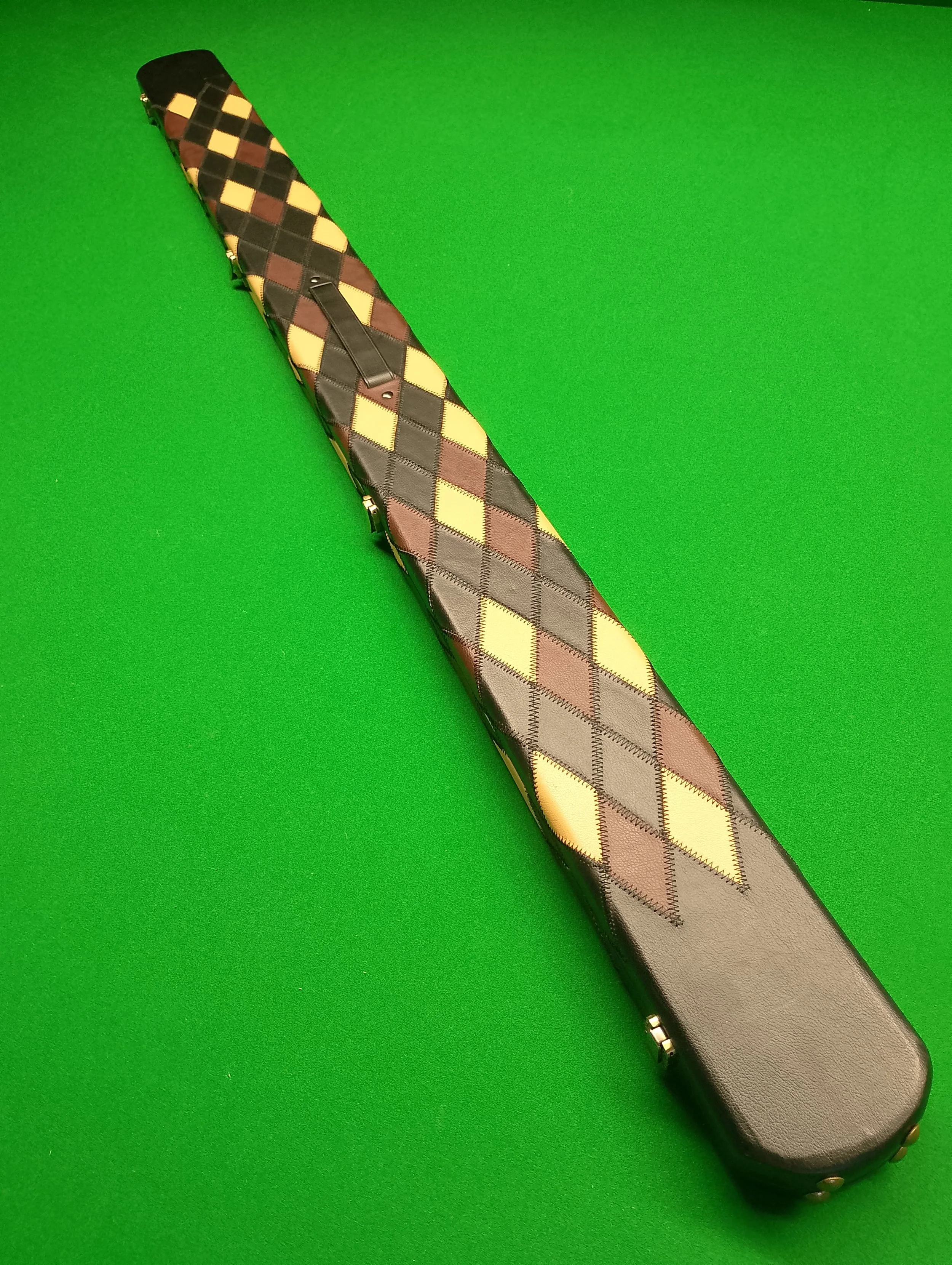 A cue stick with a quilted fabric cover in a diamond pattern of black, brown, yellow, and beige, lying on a green pool table felt.