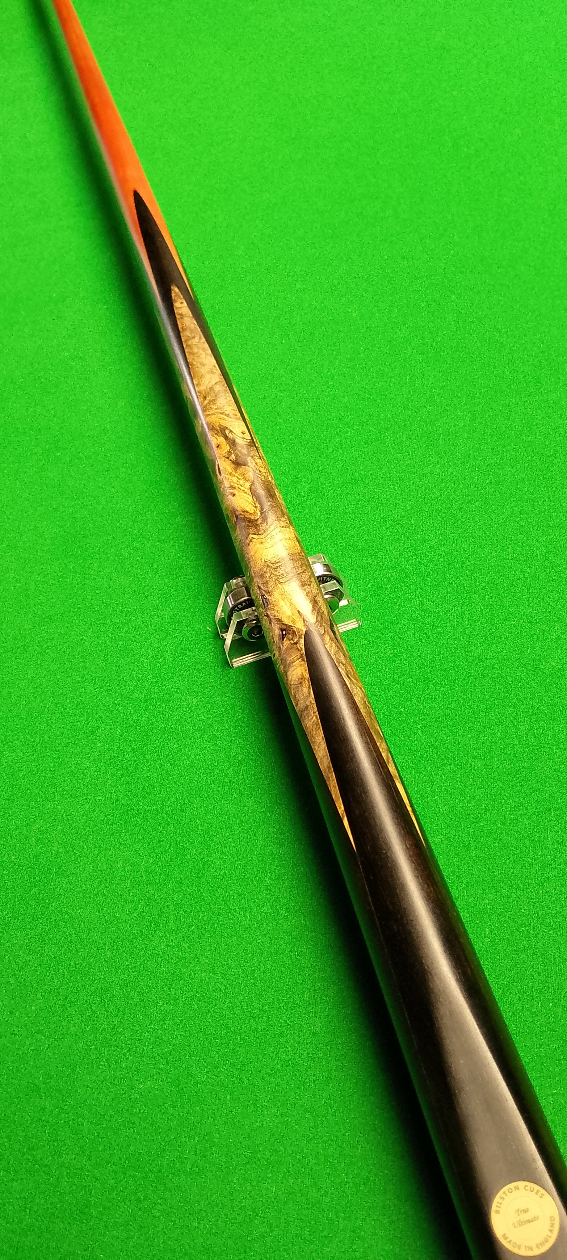 A pool cue with a black tip, a wooden body with a marbled pattern, and a label at the end that reads 'BILSTON CUES, Made in England' on a green billiard table.