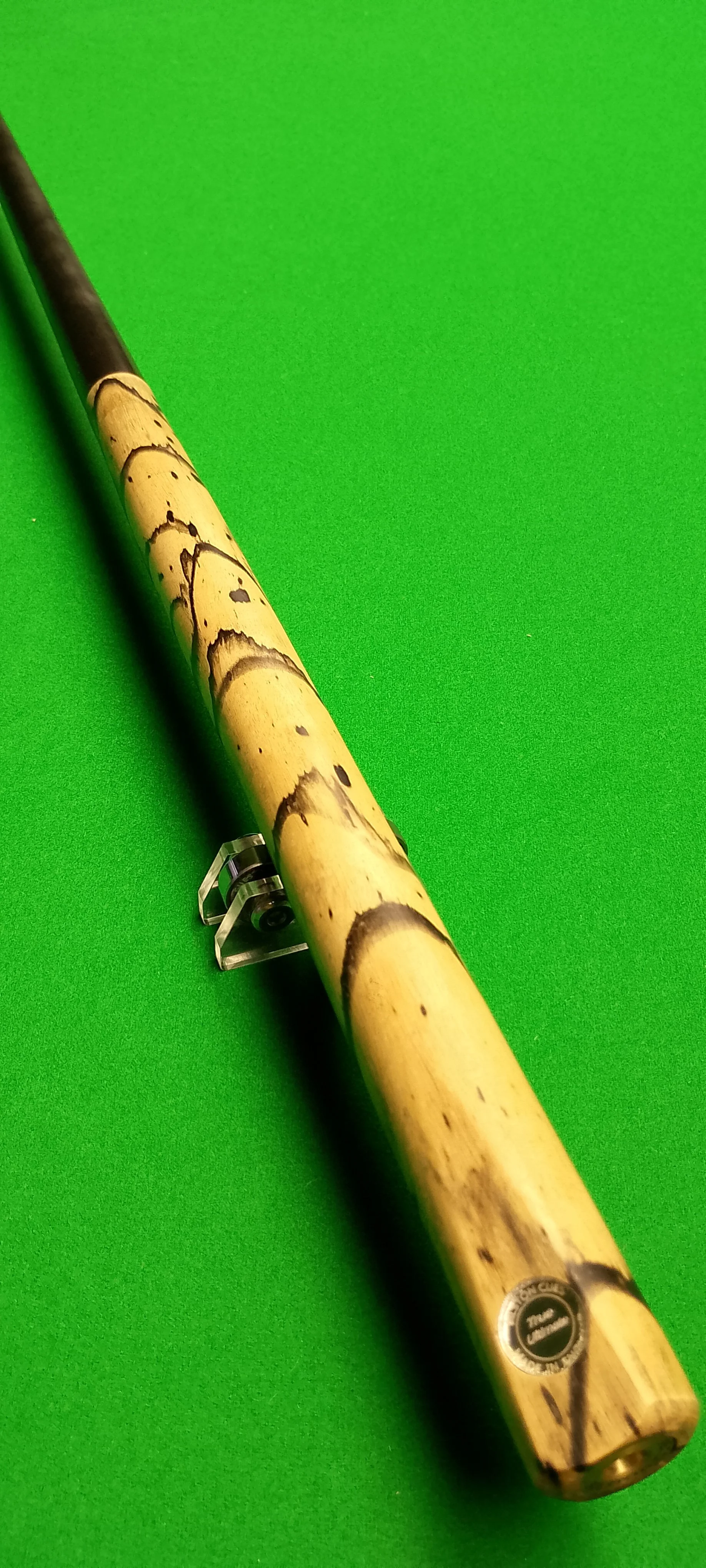 A pool cue with a beige, dark brown, and black design on a green pool table, resting on a cue stand.