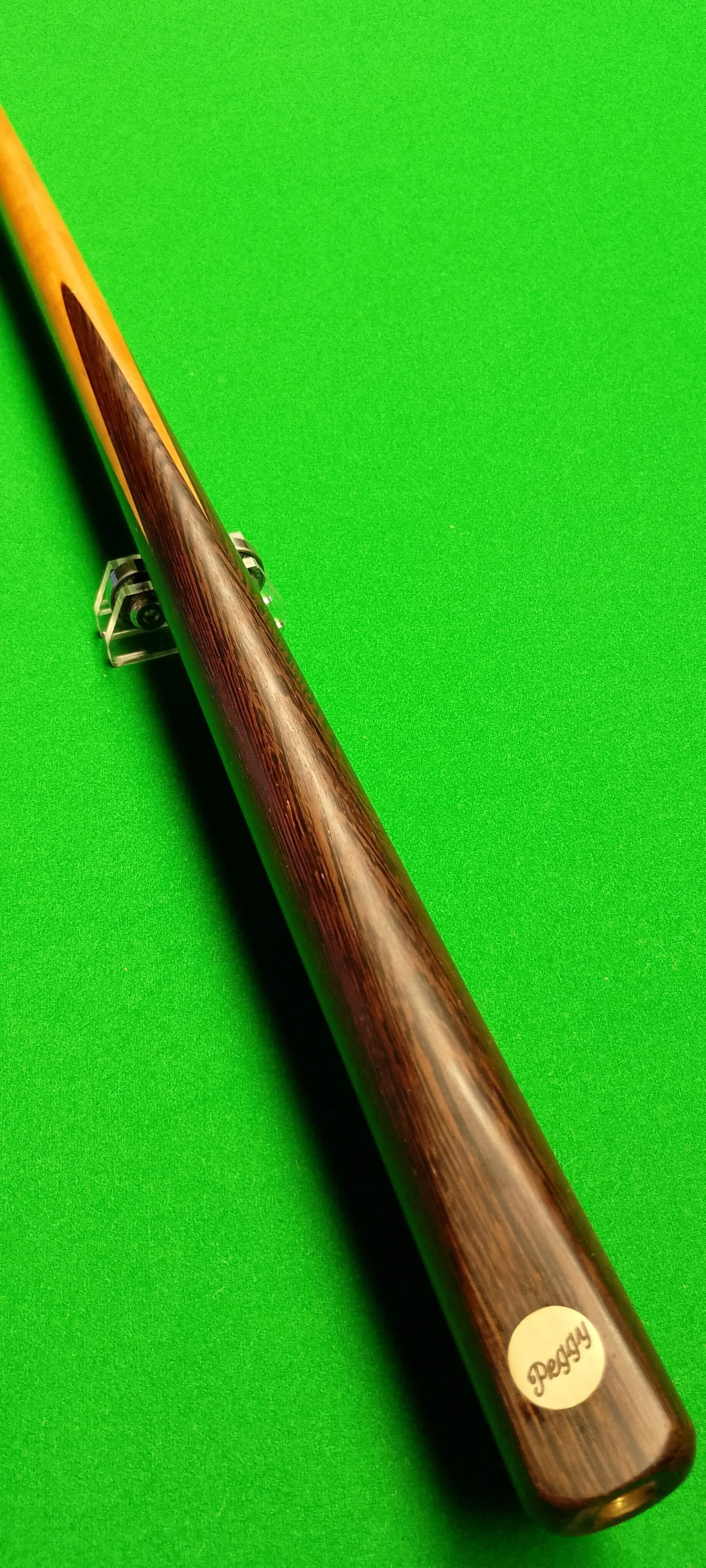 A wooden cue stick for billiards or pool placed on a green felt surface, with a small sticker near the bottom that says 'Peggy'.