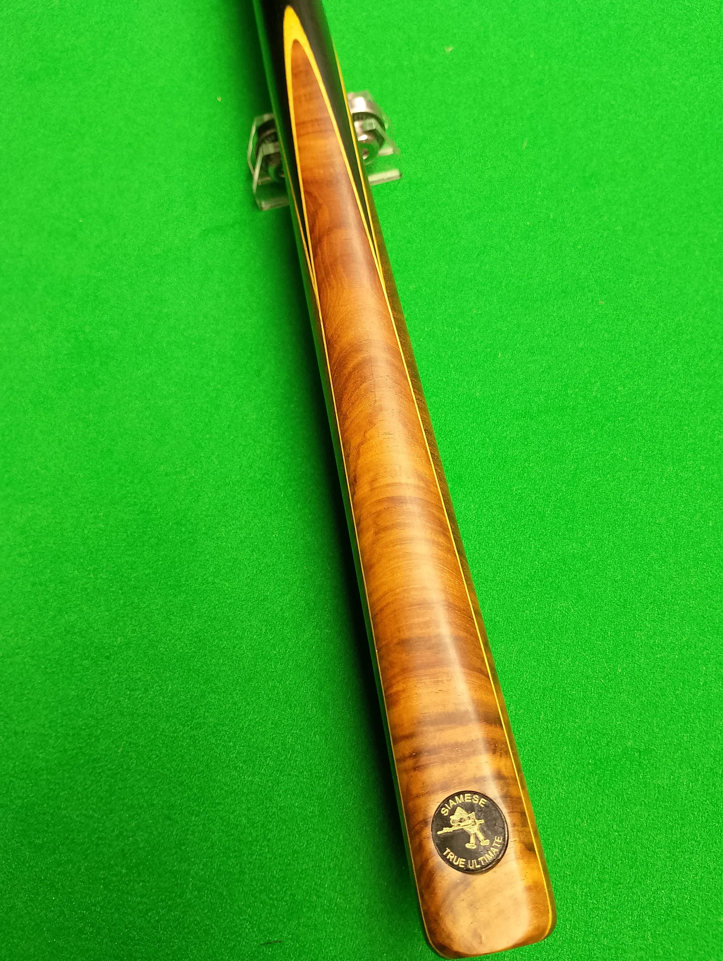 A pool cue with a wooden finish and a circular sticker at the bottom that reads 'Siamese True Ultimate,' resting on a green pool table surface.