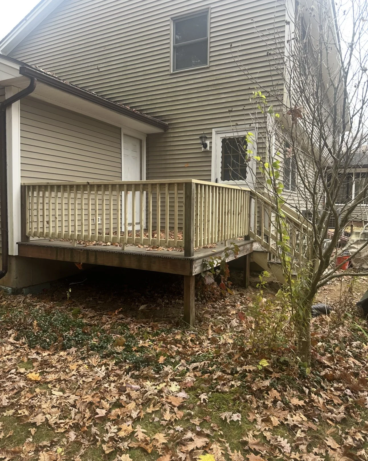 A newly built wooden deck with railings attached to the back of a house, surrounded by fallen leaves and a leafless tree in the yard.