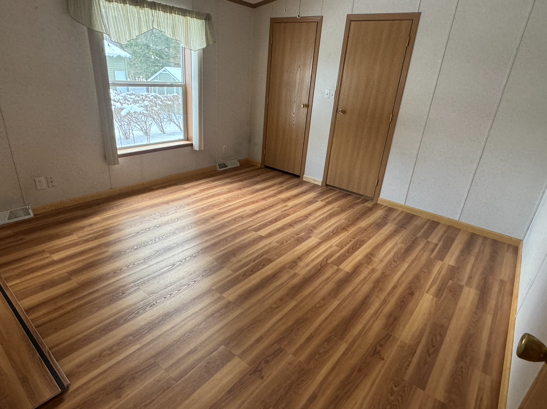 Empty room with wood flooring, a window with a green valance, two closed wooden doors, and a heater vent.