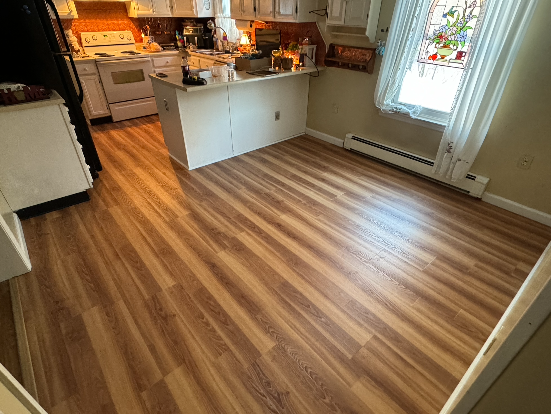 Empty kitchen and dining area with hardwood floors, white cabinets, a white stove, a kitchen island, a window with a stained glass design, and sheer curtains.
