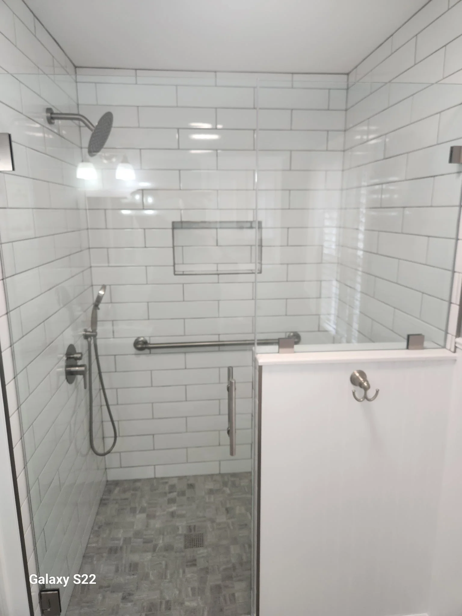 A walk-in shower with white subway tile walls, a rain showerhead, a handheld shower, a narrow glass door with a metal handle, a wall-mounted grab bar, a small wall-mounted shelf, and a dark gray tiled floor with a drain.