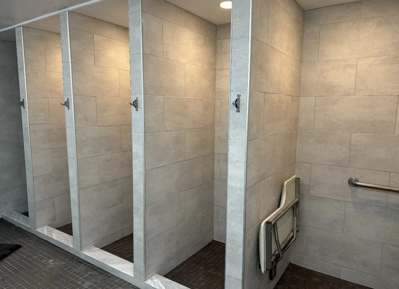 Empty shower stalls with hooks in a public bathroom, foldable chairs stored in the corner, tiled walls, and a grab bar.