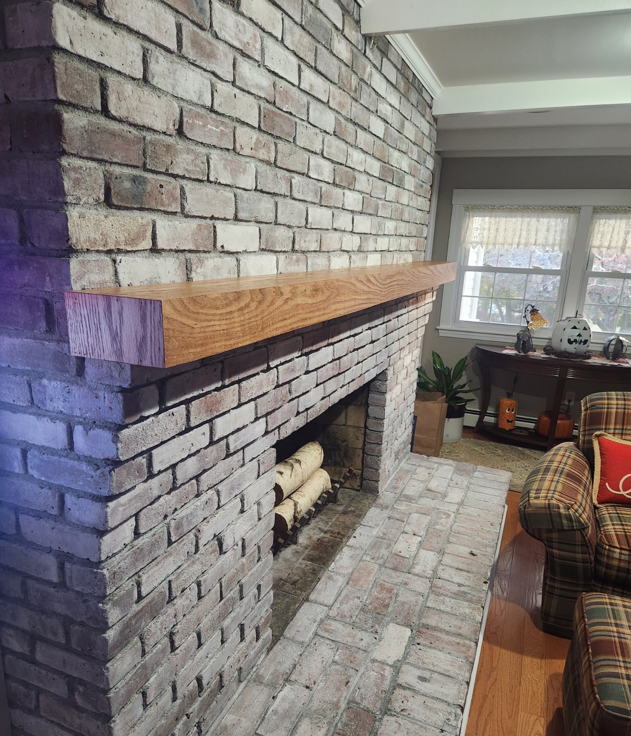 Interior of a house featuring a brick fireplace with a wooden mantel, a window with curtains, a plaid armchair, and a patio table with decorations.