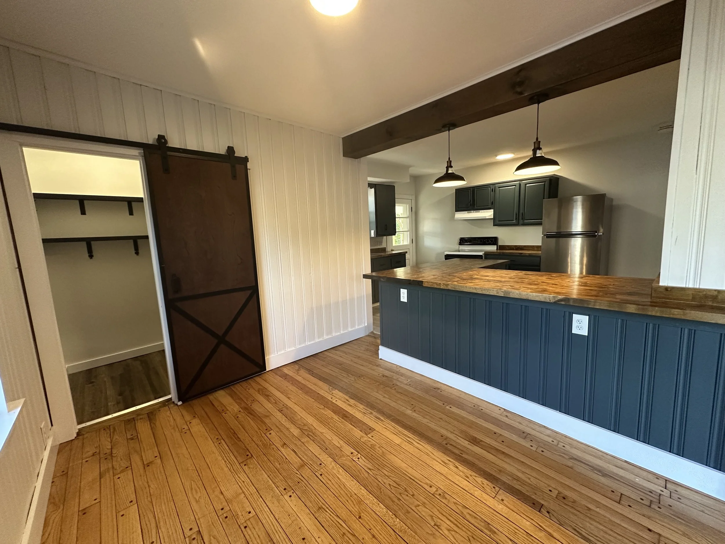 Interior of a kitchen with wooden floors, dark green cabinets, stainless steel refrigerator, and a wood countertop bar. There are two pendant lights hanging over the bar and a sliding barn door leading to a walk-in closet.